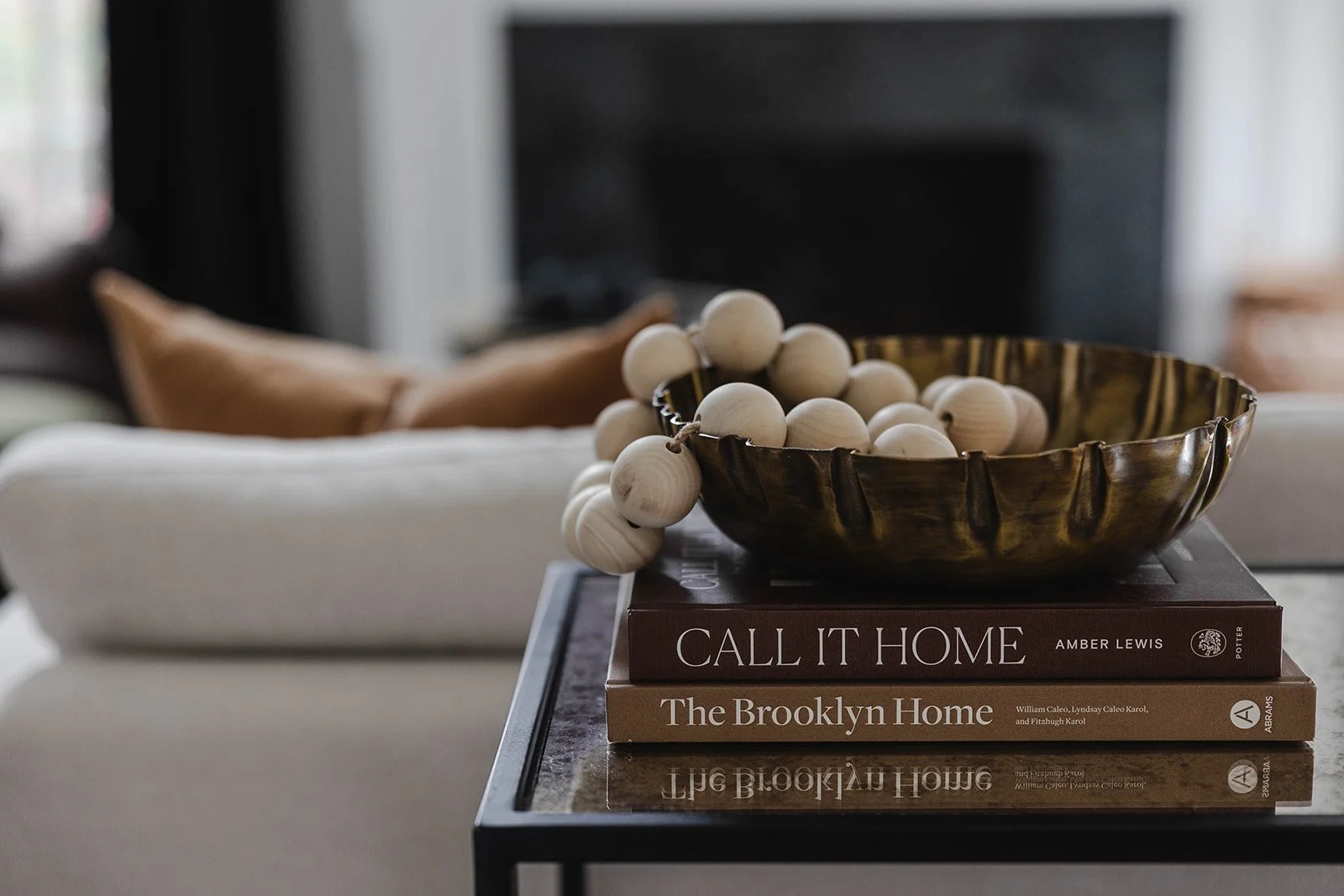 A glass bowl filled with wooden beads resting on two stacked books titled 'Call It Home' and 'The Brooklyn Home' on a glass table.