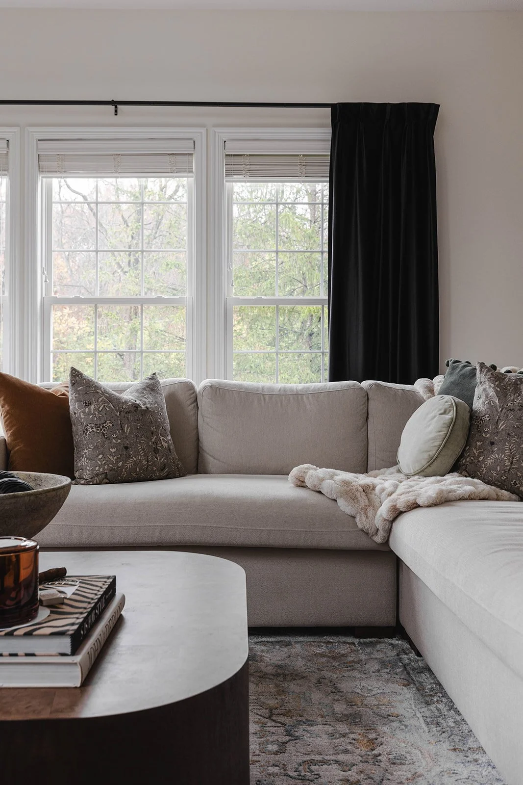 Living room with a large white sectional sofa, multiple decorative pillows, a beige fur throw blanket, a wooden coffee table with books and a candle, and large windows with white blinds and black curtains showing trees outside.