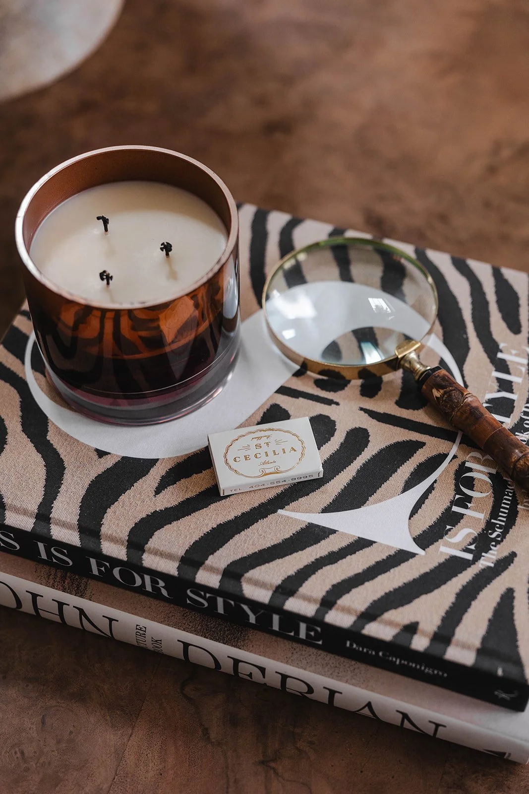 A white candle with three black wicks inside a brown glass container, placed on top of two fashion books, with a magnifying glass resting on the top book. The books are titled 'The Art of Style' and 'Diana.' There is also a small box of 'St. Cecilia'