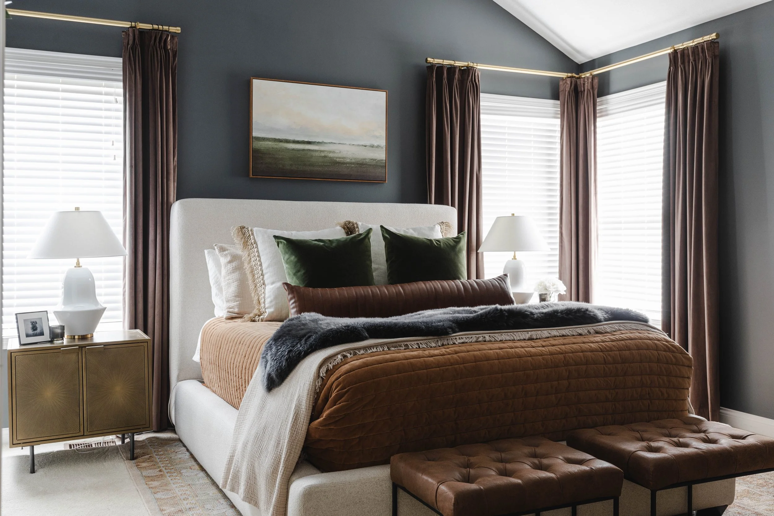 A bedroom with a white bed, dark green and beige pillows, brown and cream bedding, brown curtains, two side tables with lamps, framed painting, and two brown leather ottomans.