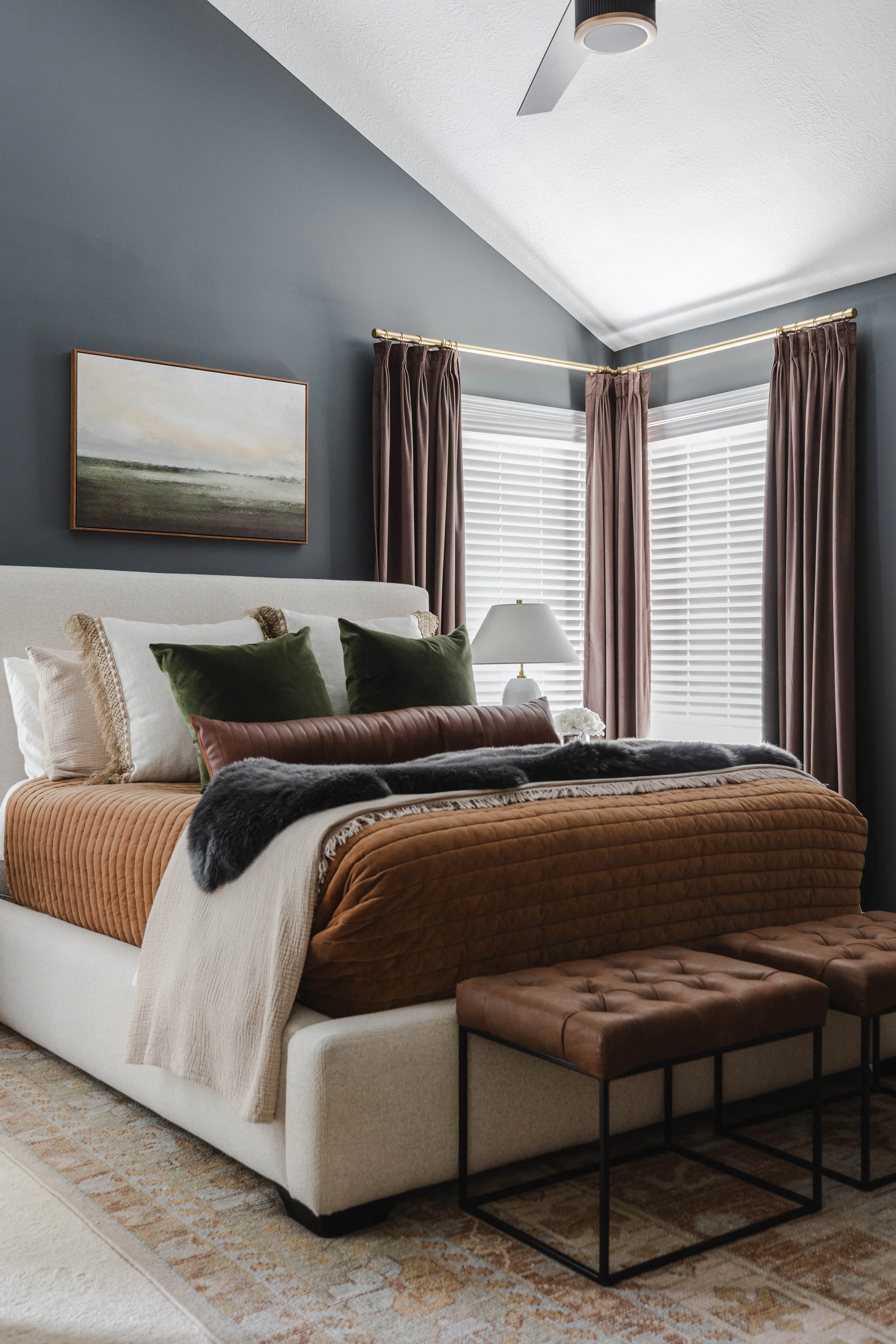 A bedroom with a beige upholstered bed, brown and green pillows, a brown bedspread, a white blanket, two brown leather stools, a white table lamp, dark brown curtains, and a landscape painting on a dark blue wall.