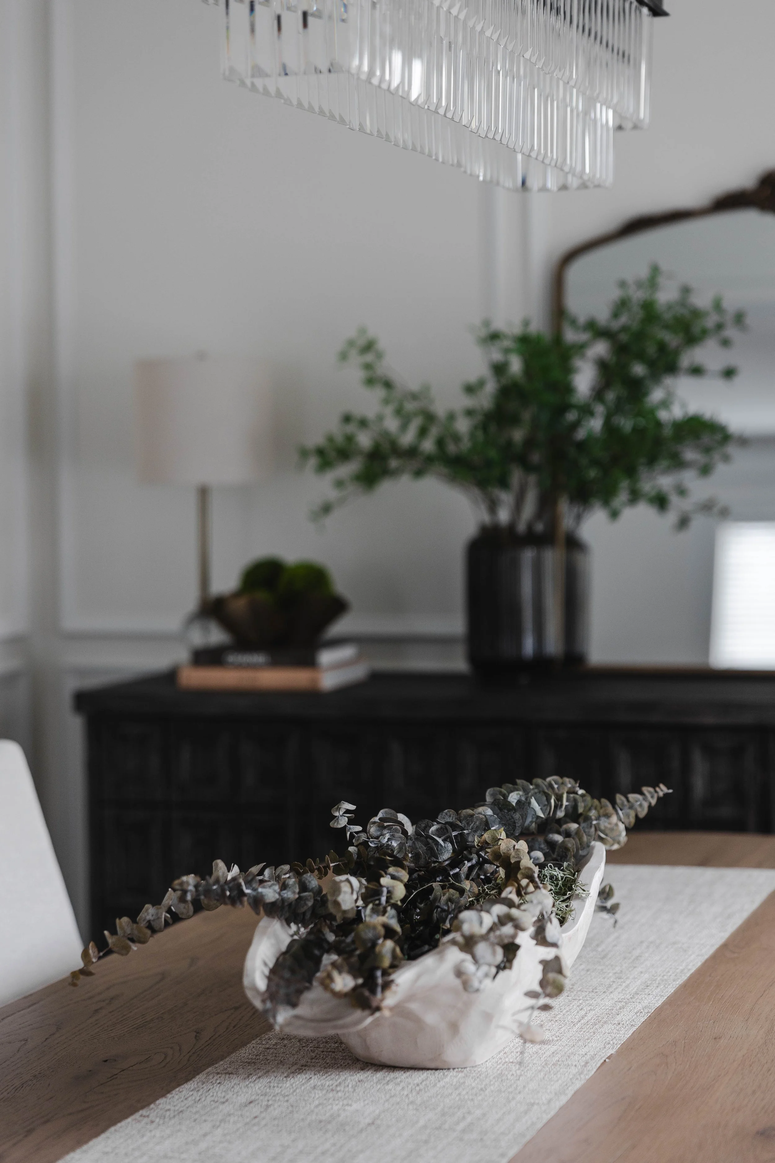 A dining room table with a white centerpiece containing dried eucalyptus branches, with a black sideboard and green foliage in a black vase in the background.