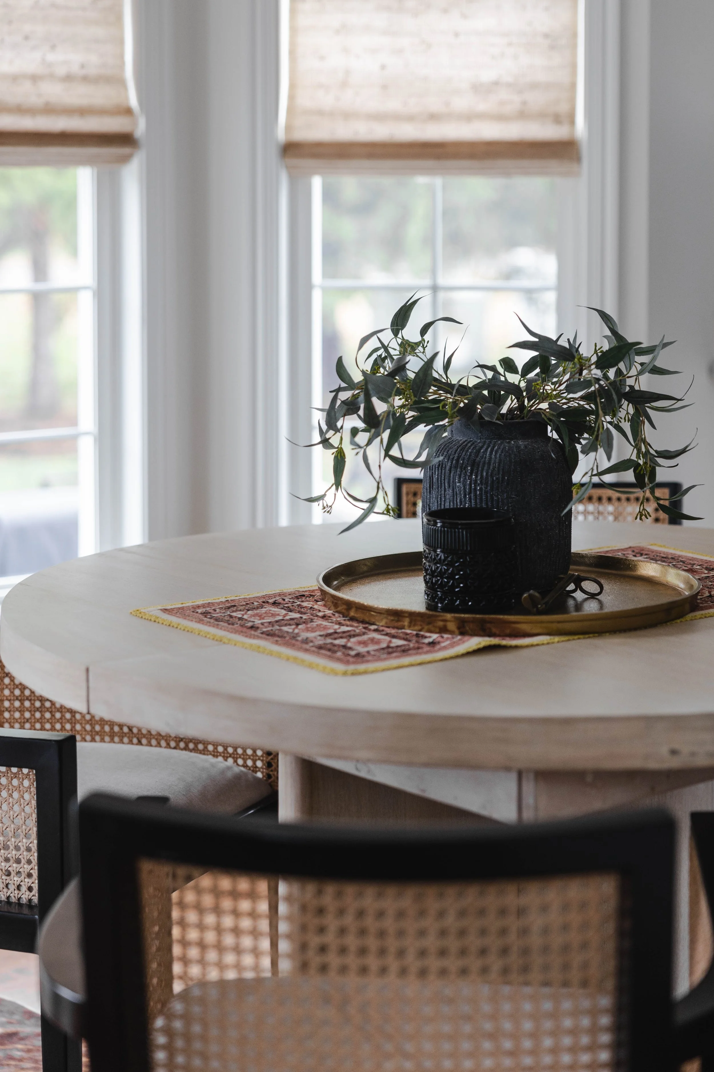A round wooden dining table with a decorative plant in a dark textured vase, set on a patterned table runner, with black glassware on a gold tray, located near windows with beige blinds.