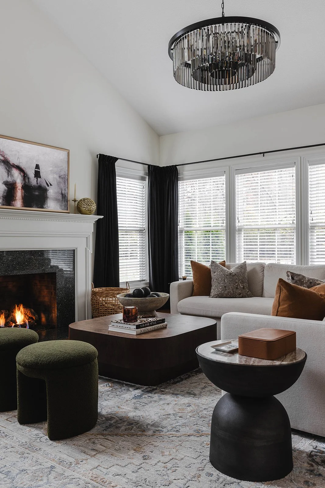 Living room with a white sofa, a black coffee table, green ottomans, a fireplace with a fire, and a large window with black curtains, a modern chandelier, and decorative pillows.