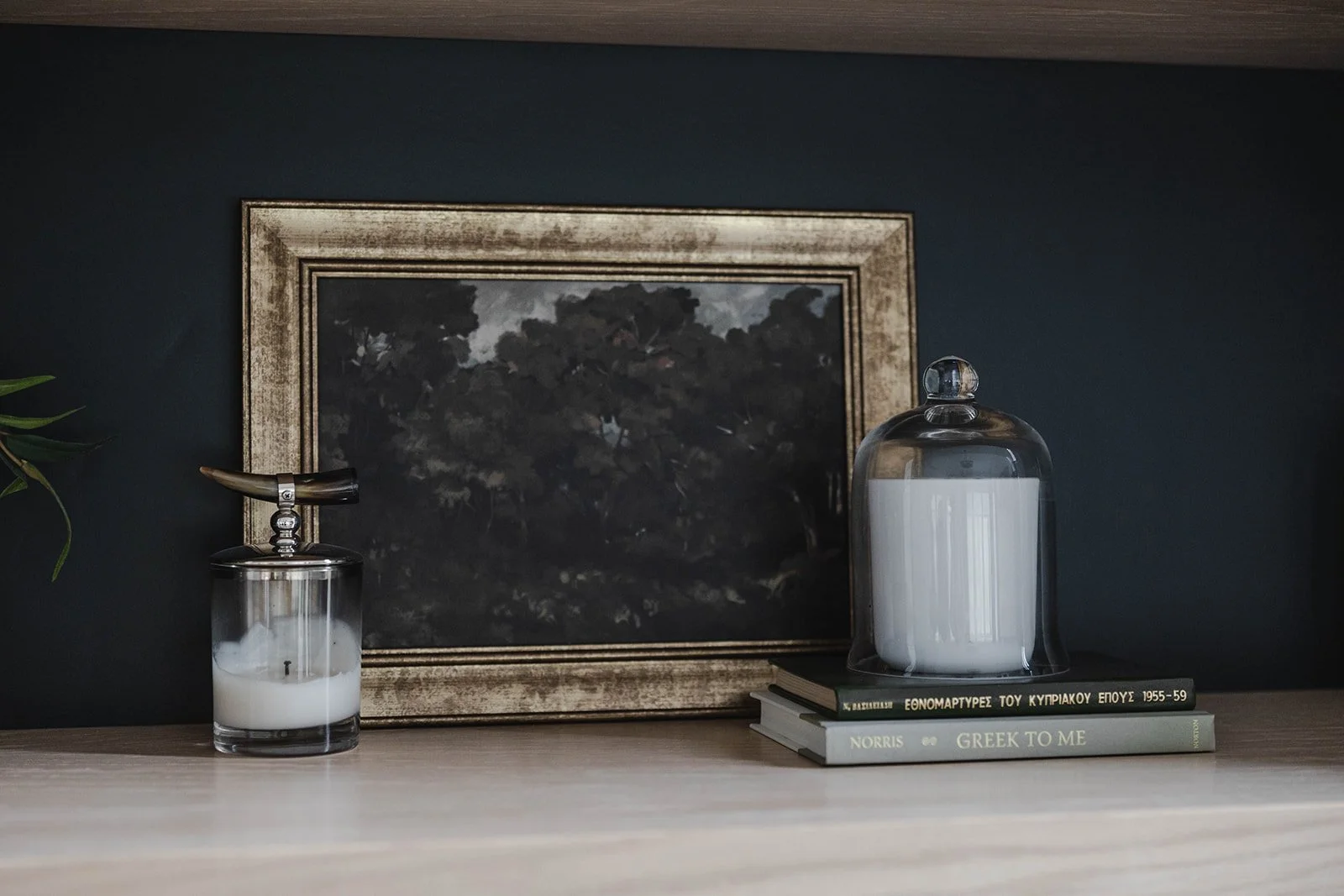 Decorative tabletop arrangement with candles, a framed painting of dark trees, and books on a wooden surface against a dark wall.