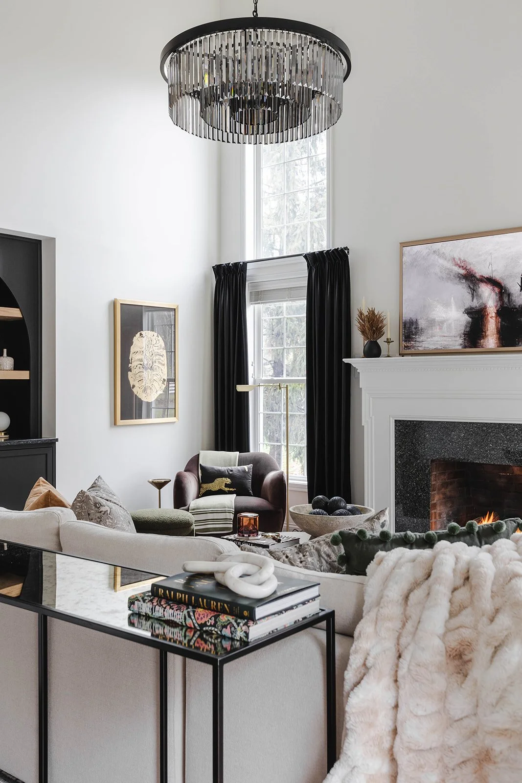A stylish living room featuring a black chandelier, white walls, large windows with black curtains, a white fireplace, and various modern decor and furniture including a beige sofa, a dark armchair, and a glass-top side table with books and decorativ