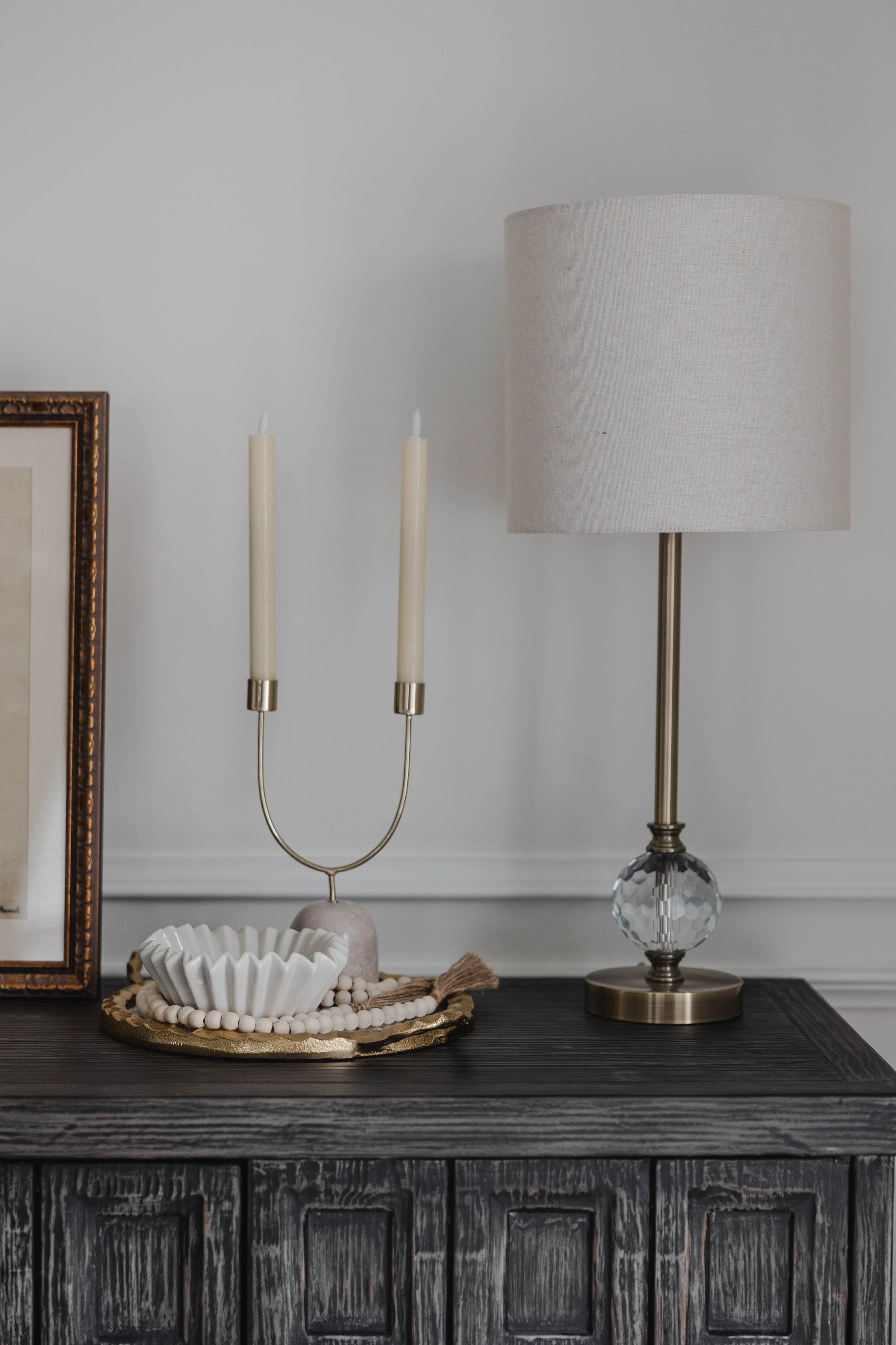 A dark wooden table with a beige lampshade on a brass and glass lamp, a gold-framed picture, and a decorative white bowl with beads, along with a beige candle holder with two white candles.