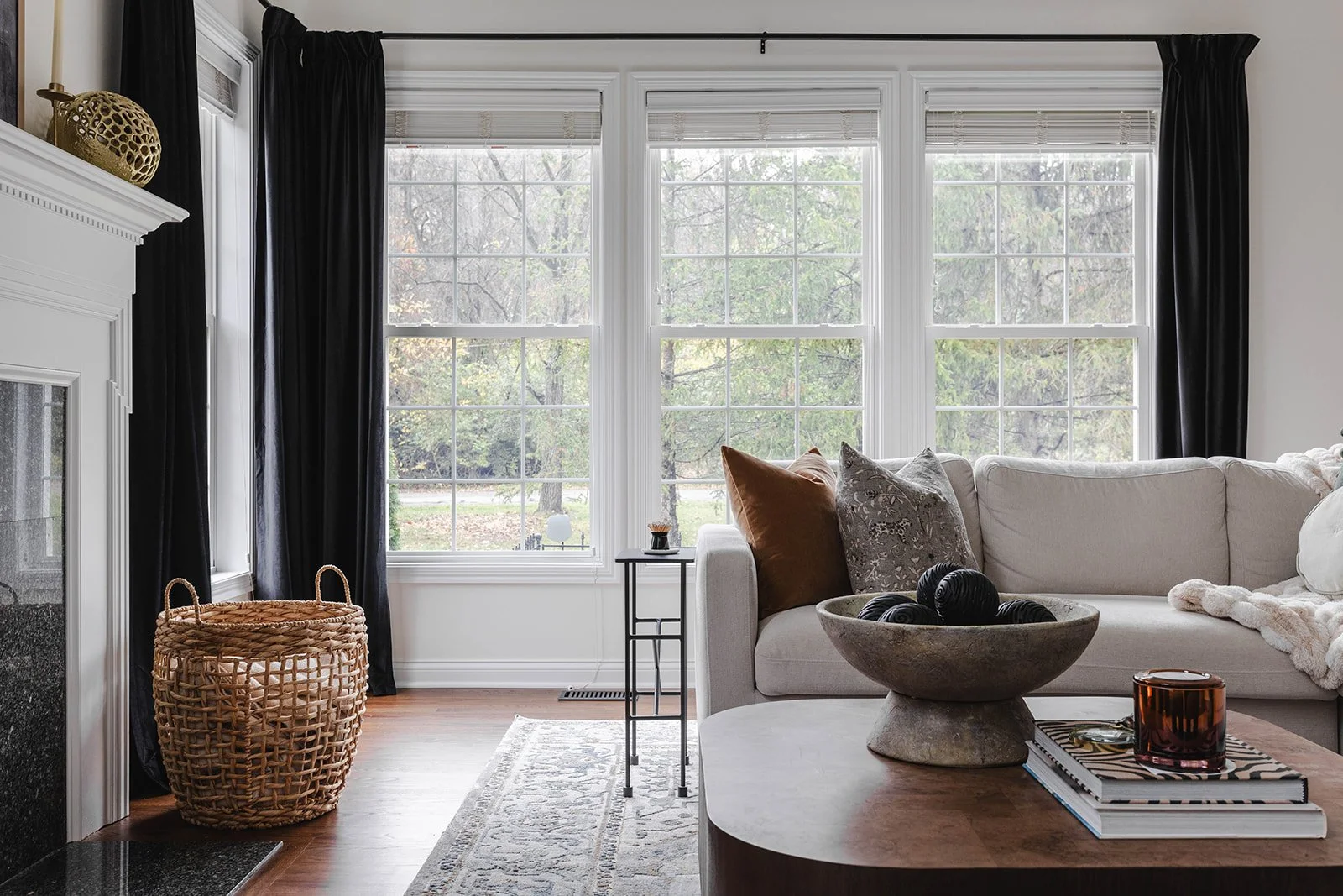 Living room with large windows, black curtains, white sofa with pillows, woven basket, fireplace, small side table, decorative bowl with black objects, books, and candles.