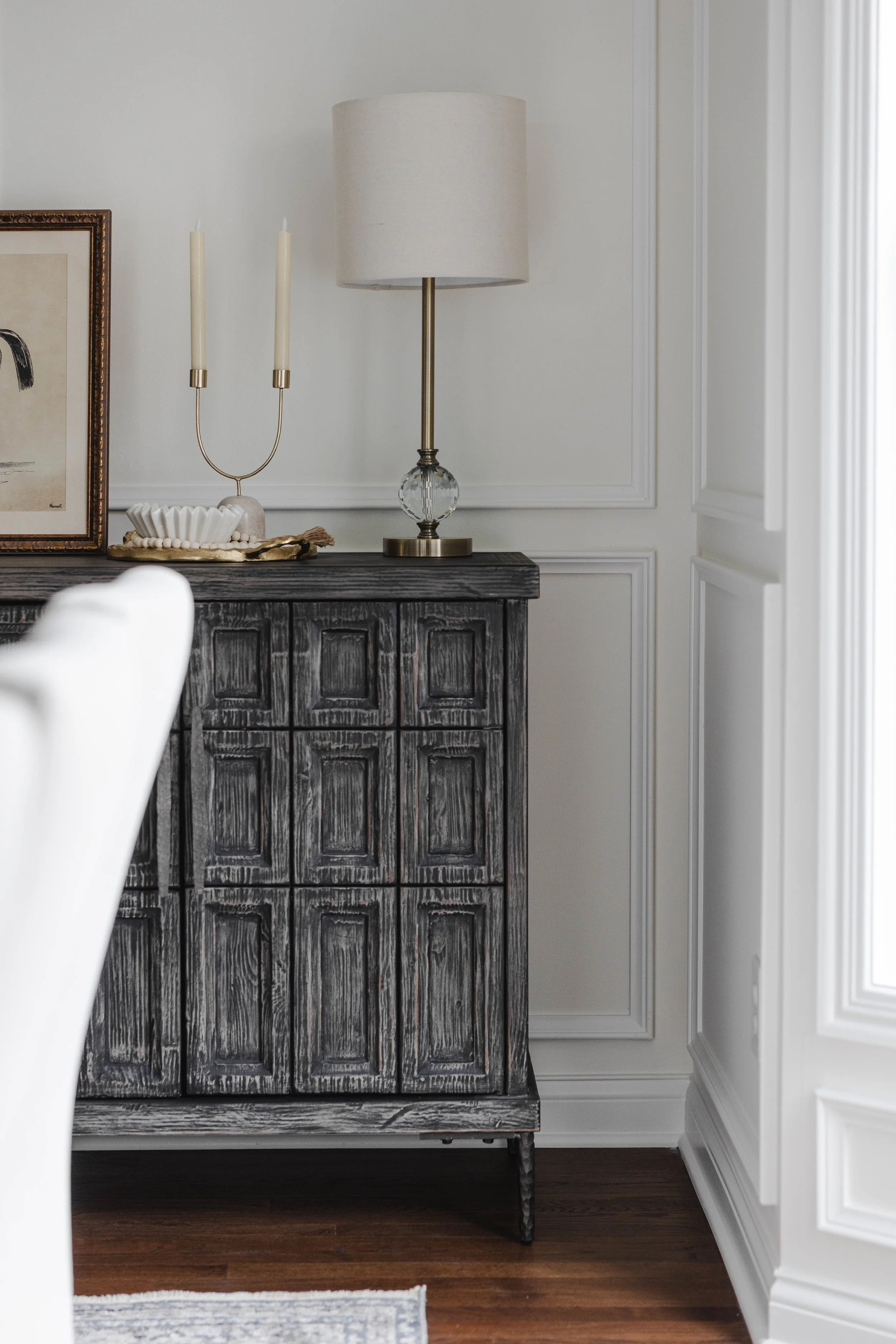 A black distressed wood sideboard with decorative paneling sits against a white wall in a room with wood flooring. On top of the sideboard is a white ceramic shell dish, a gold tray, a candle holder with two lit candles, and a table lamp with a beige
