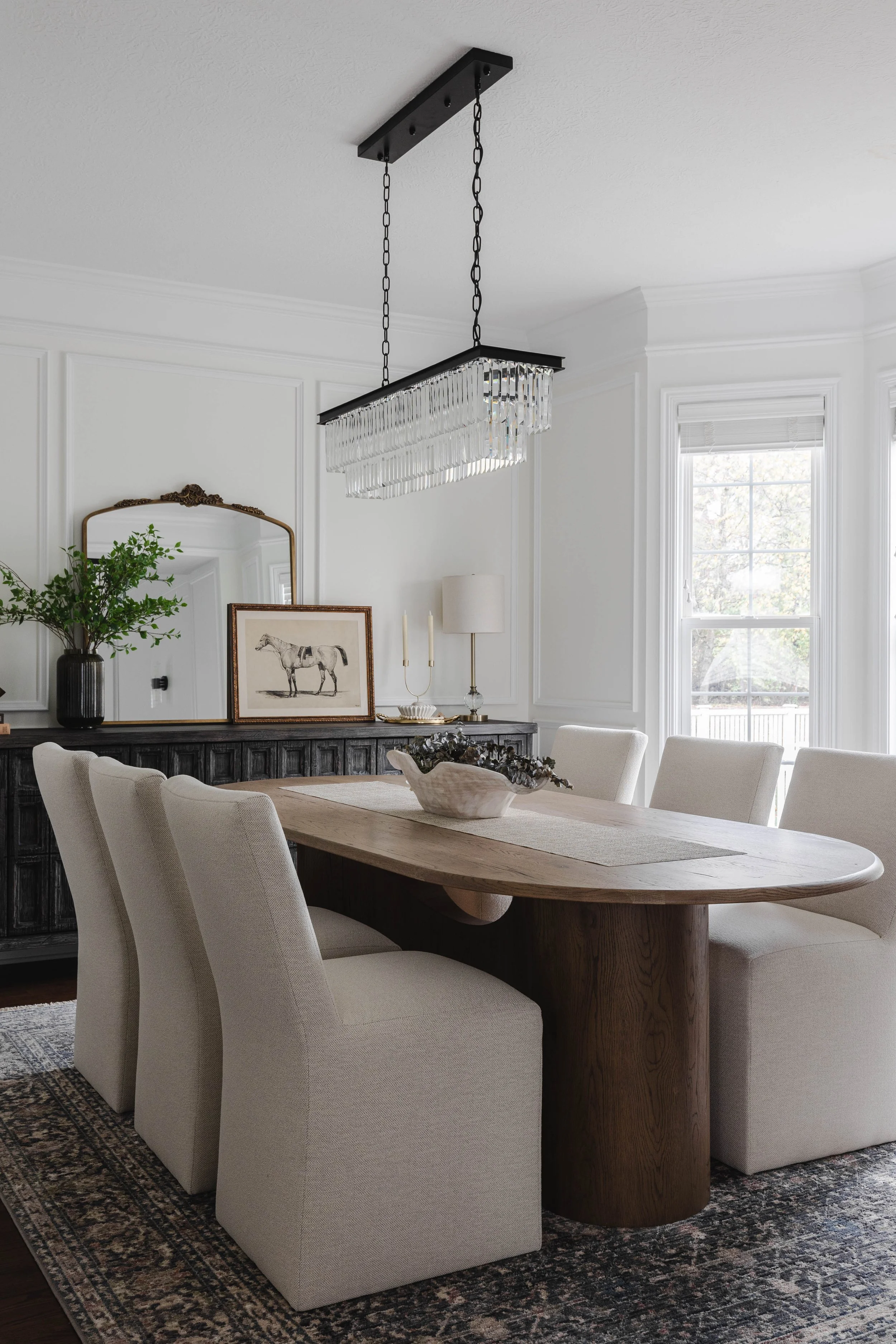 A dining room with a wooden table surrounded by six beige upholstered chairs, a black sideboard with decorative items, a mirror, and framed art, with large windows and a modern chandelier hanging from the ceiling.