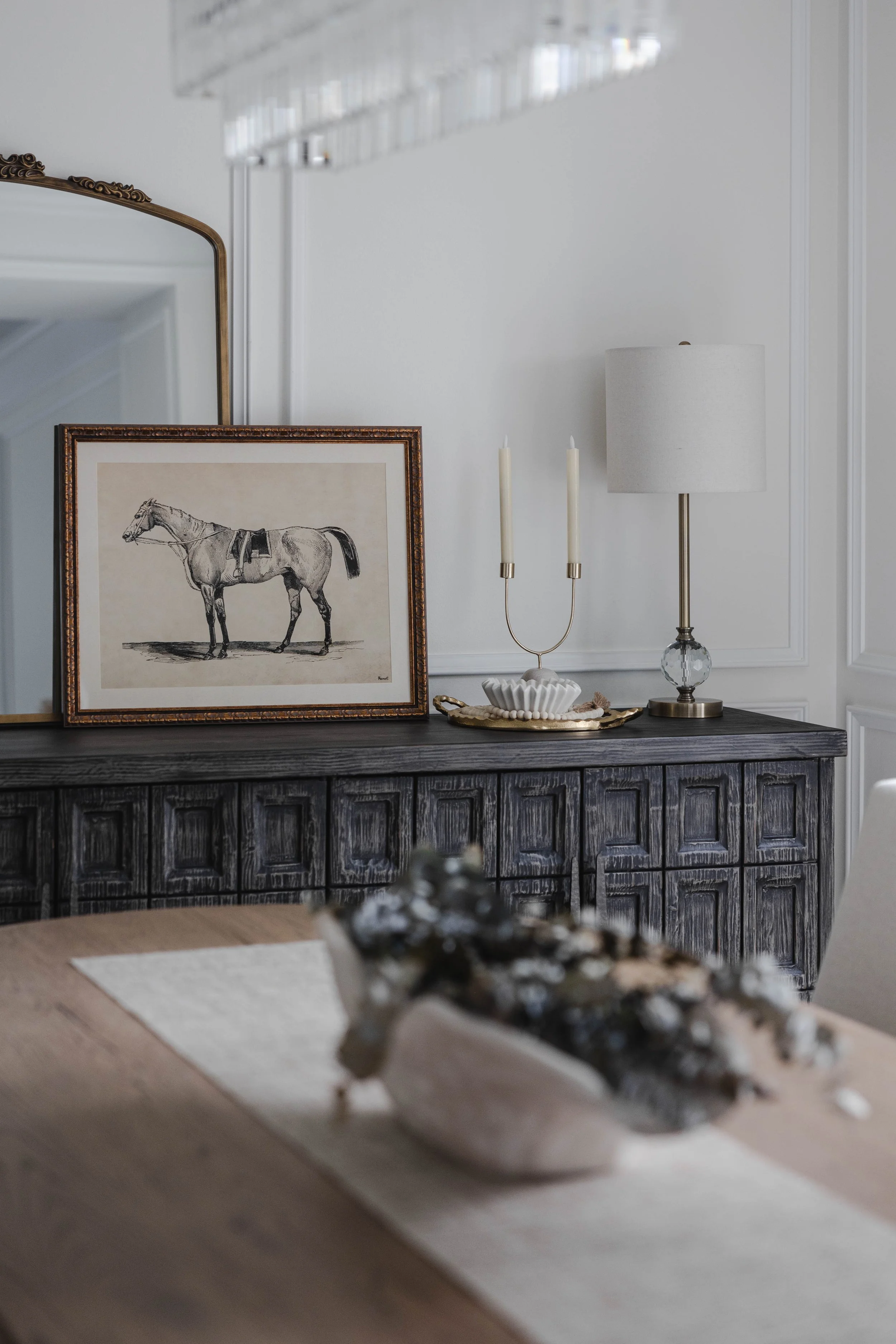 A sideboard with a framed horse drawing, white candlesticks, a table lamp, and platter in a room with white walls and trim.