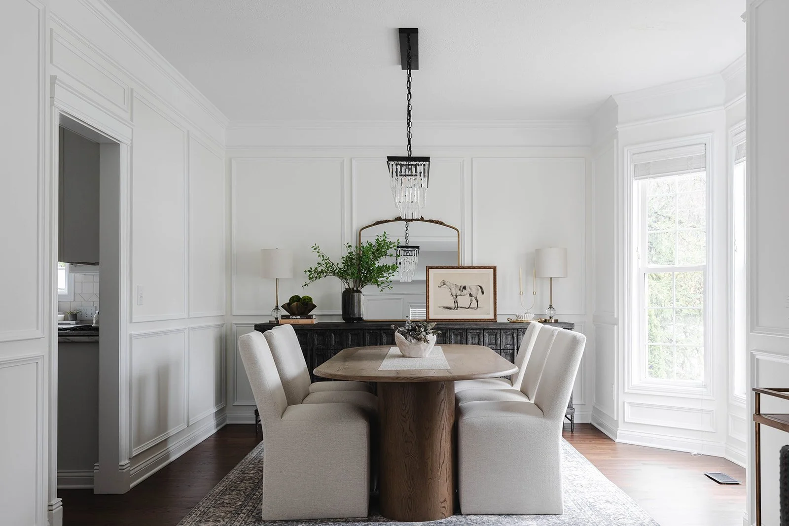 Elegant dining room with a wooden table surrounded by six cream-colored upholstered chairs, a black sideboard with decorative items, two white lamps, a framed horse illustration, and a large window letting in natural light.