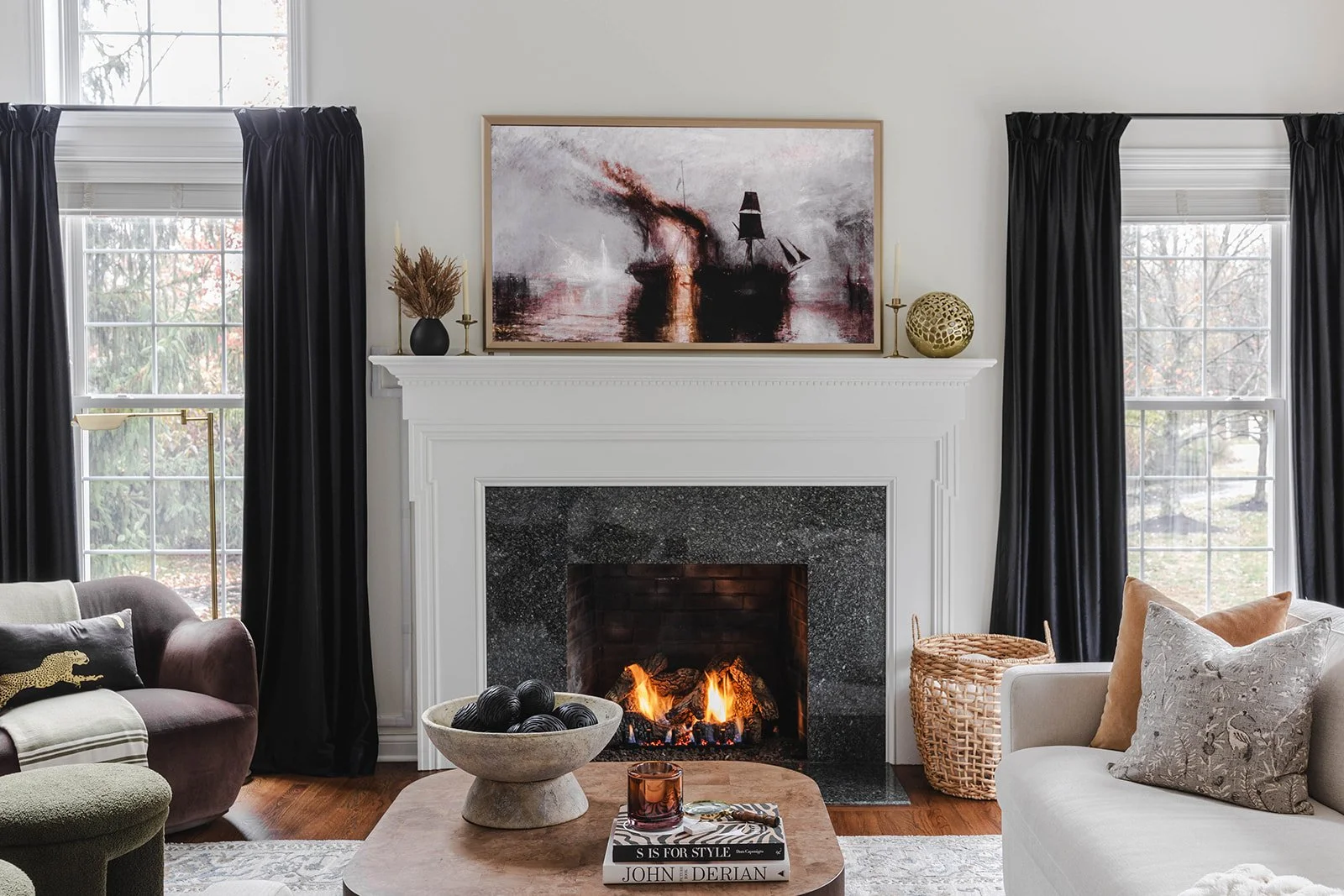 A cozy living room with a white fireplace, lit with a fire, flanked by two windows with black curtains. Above the fireplace hangs a large abstract painting of a ship in stormy seas. Decor items on the mantel include a black vase with dried plants, ca