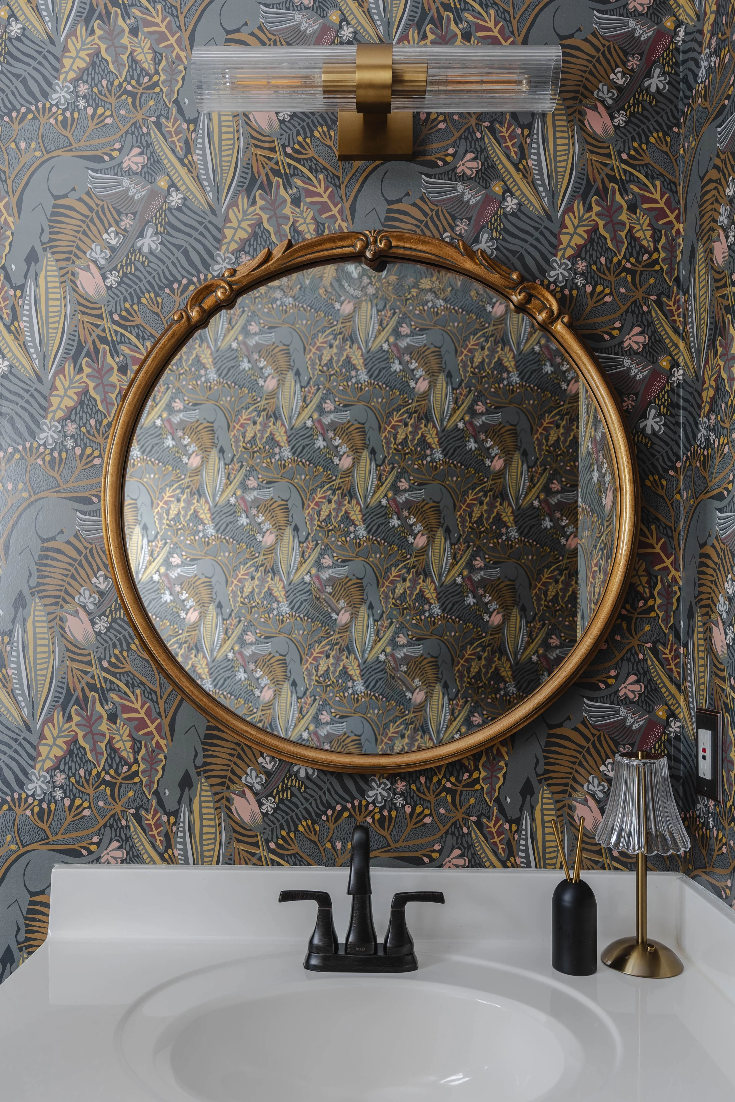 A bathroom vanity with a white sink, black faucet, and decorative items. Above the sink is a round mirror with a gold frame, and above the mirror is a modern wall-mounted light fixture with a gold base and a clear glass shade. The wall has a colorful