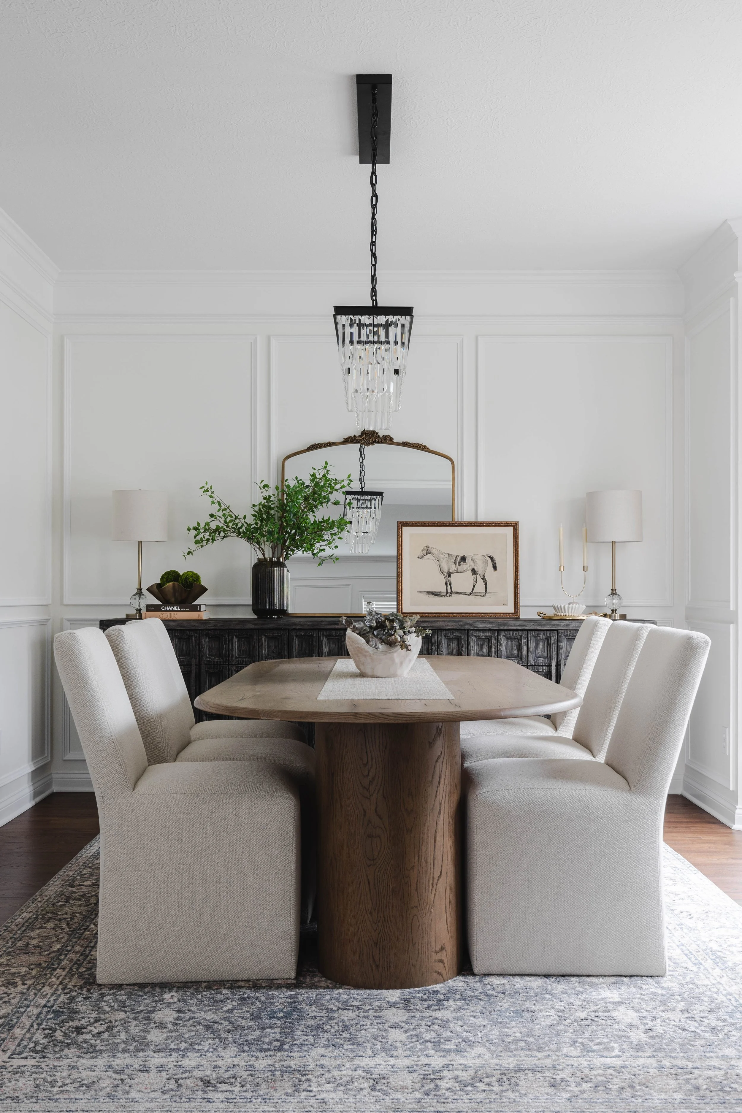 Dining room with a wooden table, six upholstered chairs, a black sideboard with decorative items, a large mirror, framed horse artwork, a chandelier, and table lamps.