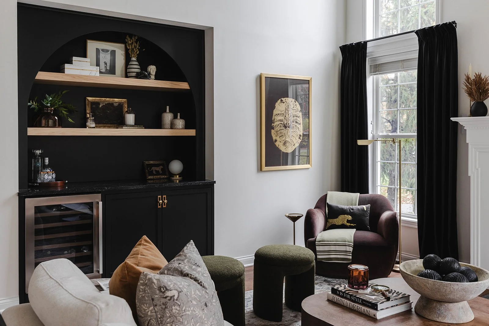 Living room with white walls, black curtains, a window, a maroon armchair with cushions, a round coffee table with books and decorative objects, and built-in shelves with decorative items.