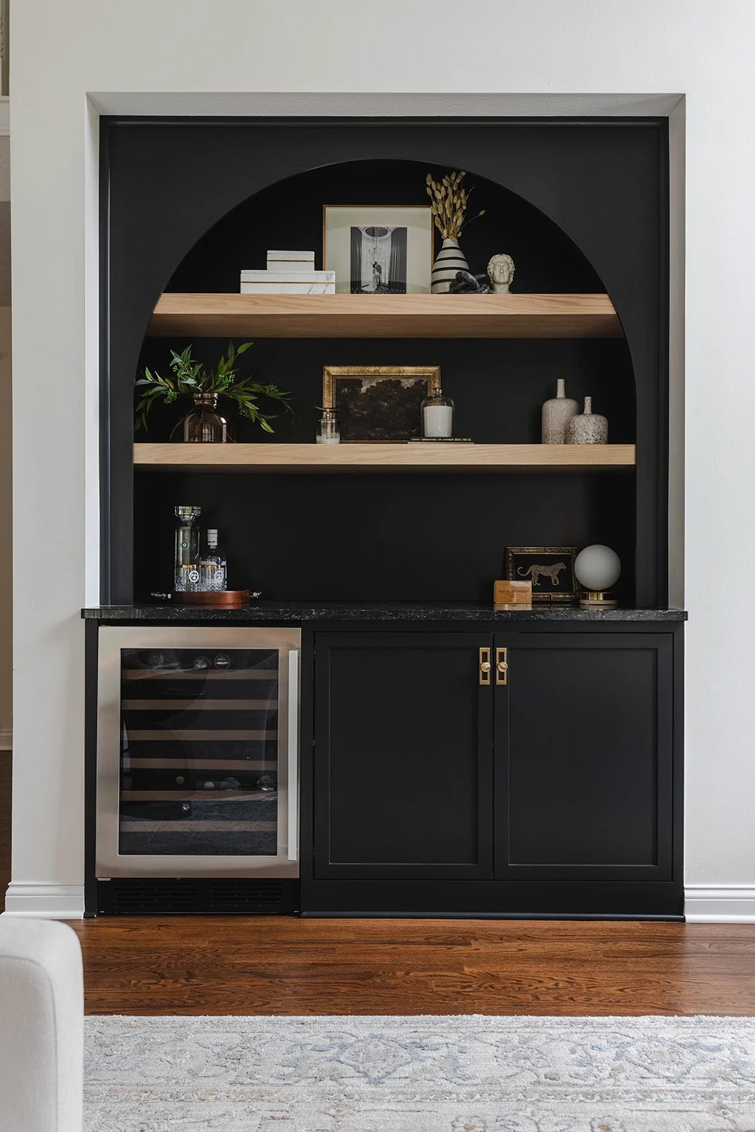 Built-in black and wood bar cabinet with shelves, bottles, decor items, and a mini wine fridge under a marble countertop.