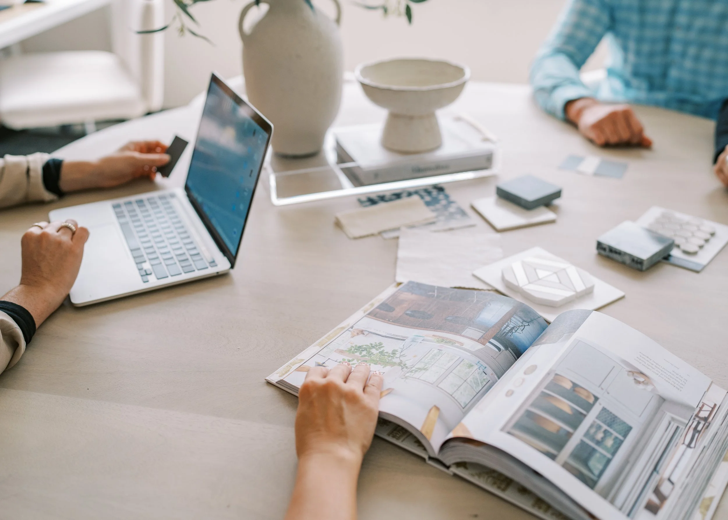 A group of people sitting around a table working on a project, with a laptop, a magazine, and various design samples.