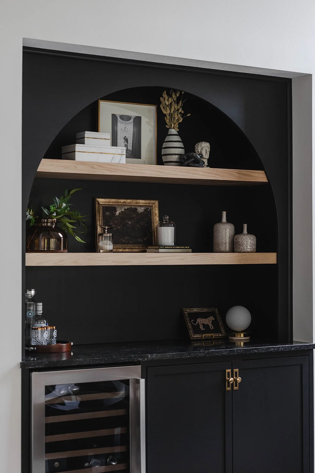 Built-in black bookshelf with three wooden shelves decorated with framed pictures, vases, sculptures, and books, installed above a black countertop with a wine cooler to the left.