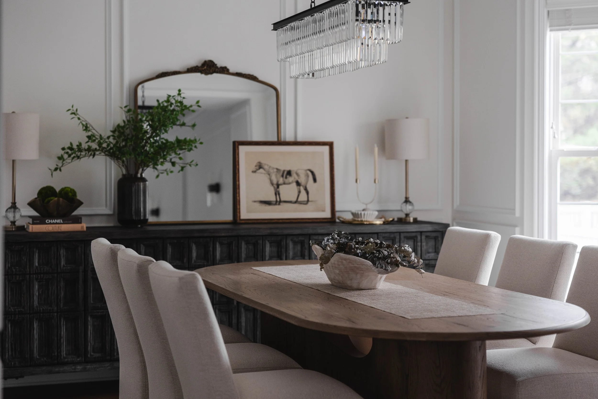 A dining room with a wooden table surrounded by beige upholstered chairs, a black sideboard with decorative objects including a framed horse print, a green plant in a black vase, and a mirror, with a window letting in natural light.