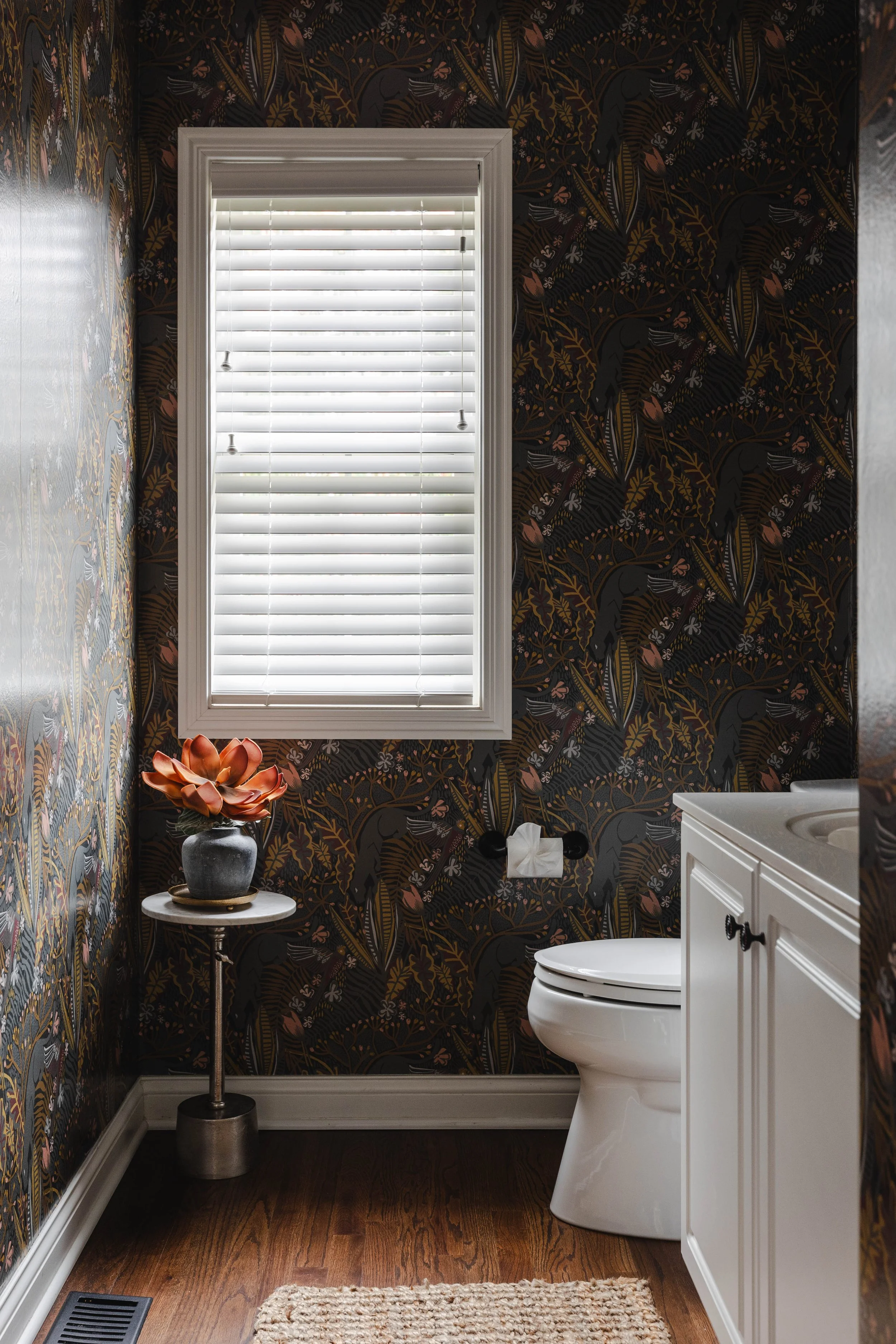 A small bathroom with dark floral wallpaper, a white window blind, a toilet, a white cabinet, and a small side table with a plant in a vase.