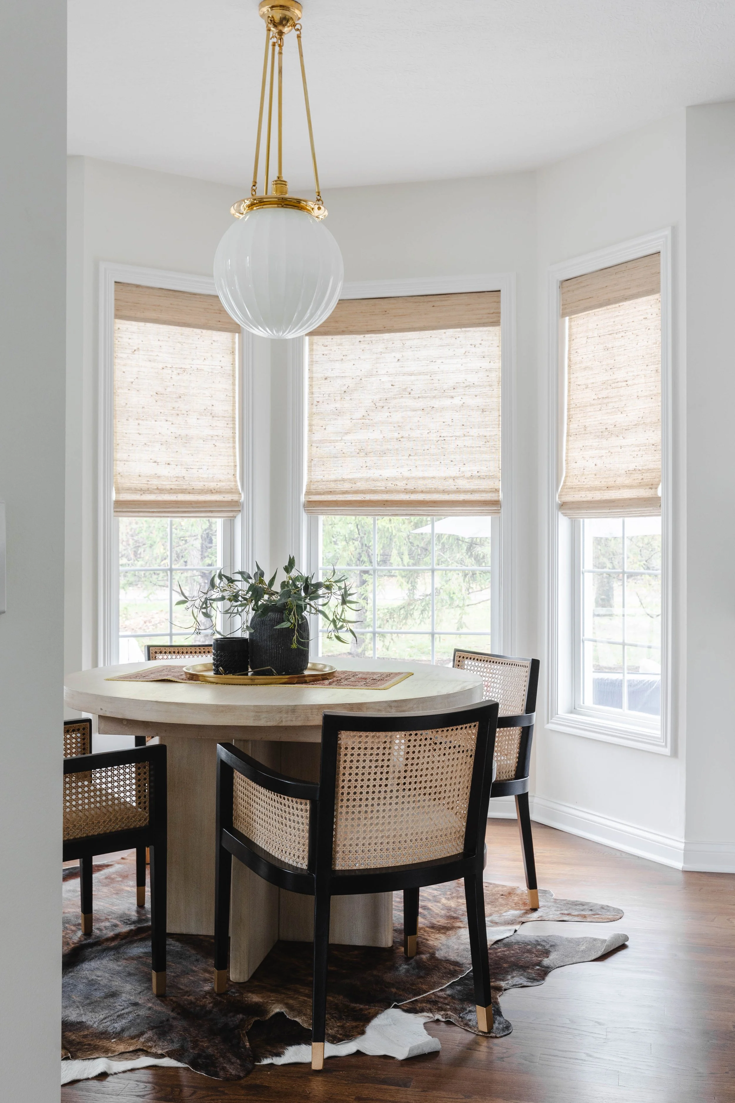 A bright dining room with a round table, four black framed chairs with woven seats, a cowhide rug, large windows with beige blinds, and a hanging globe light fixture.