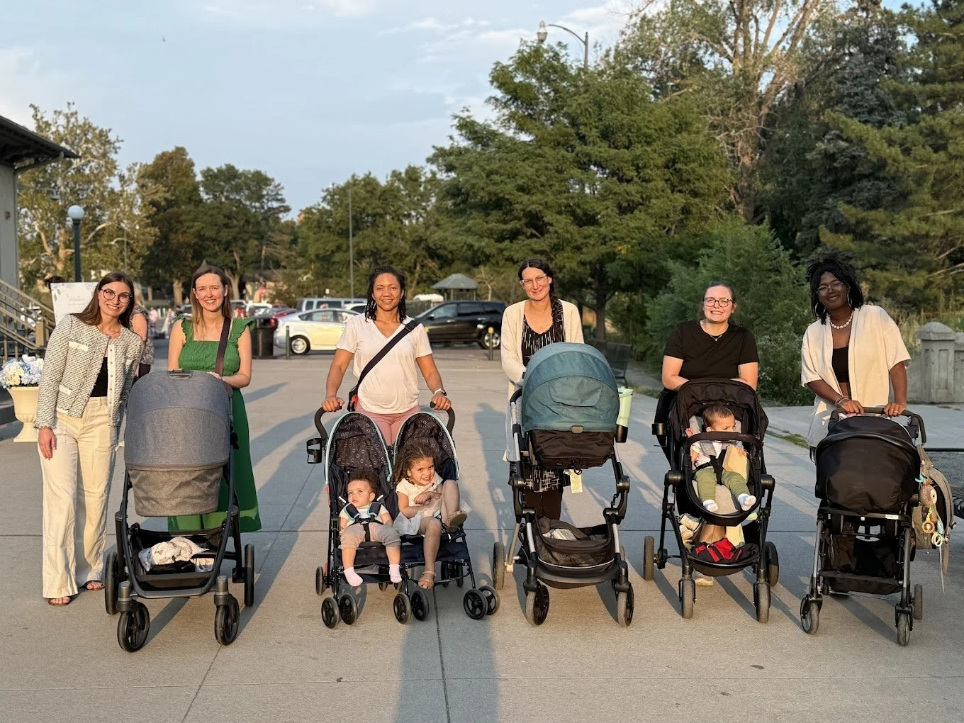 Group of six women and four children with strollers standing outdoors on a sidewalk with trees and parked cars in the background during the day.