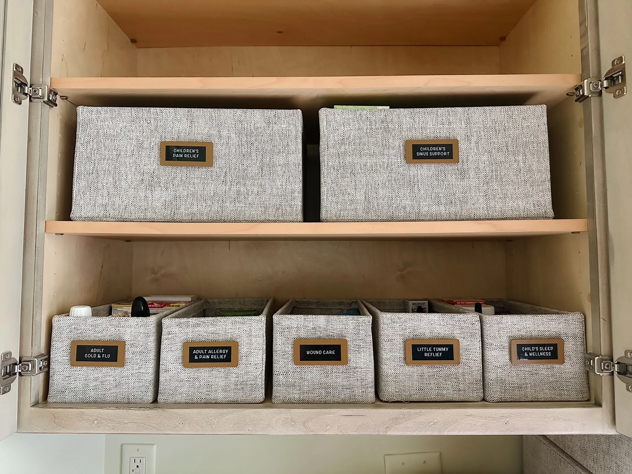 A wooden cabinet with six fabric-lined labeled storage bins. The top row has two bins labeled 'Children's Pain Relief' and 'Children's Sinus Support.' The bottom row has four bins labeled 'Adult Cold & Flu,' 'Adult Allergy & Pain Relief,' 'Wound Care,' 'Little Tummy Relief,' and 'Child's Sleep & Wellness.'