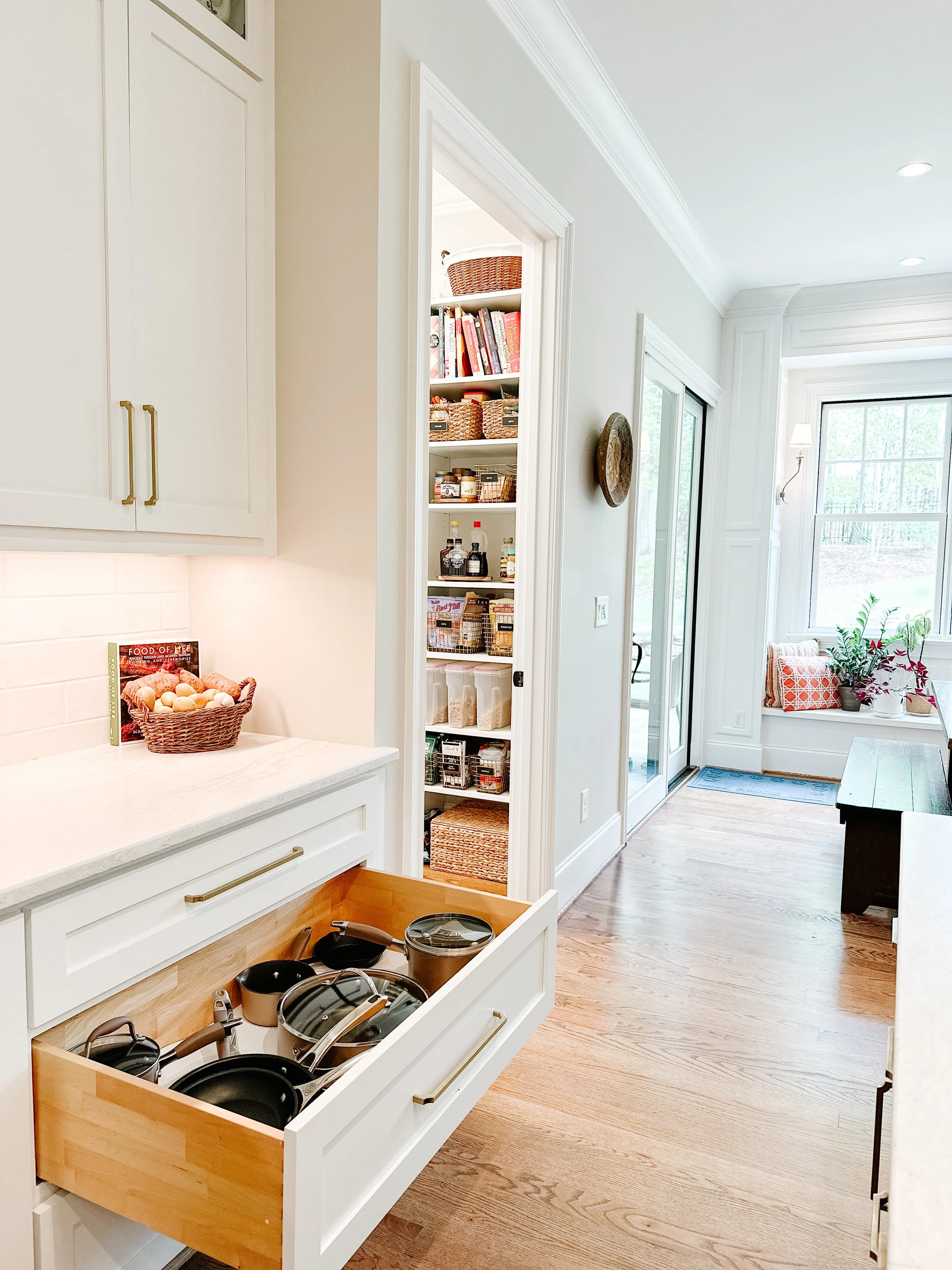 A kitchen with white cabinets, a drawer open showing cookware, a small pantry with shelves full of jars and baskets, a sliding glass door, and a window seat with cushions and plants.