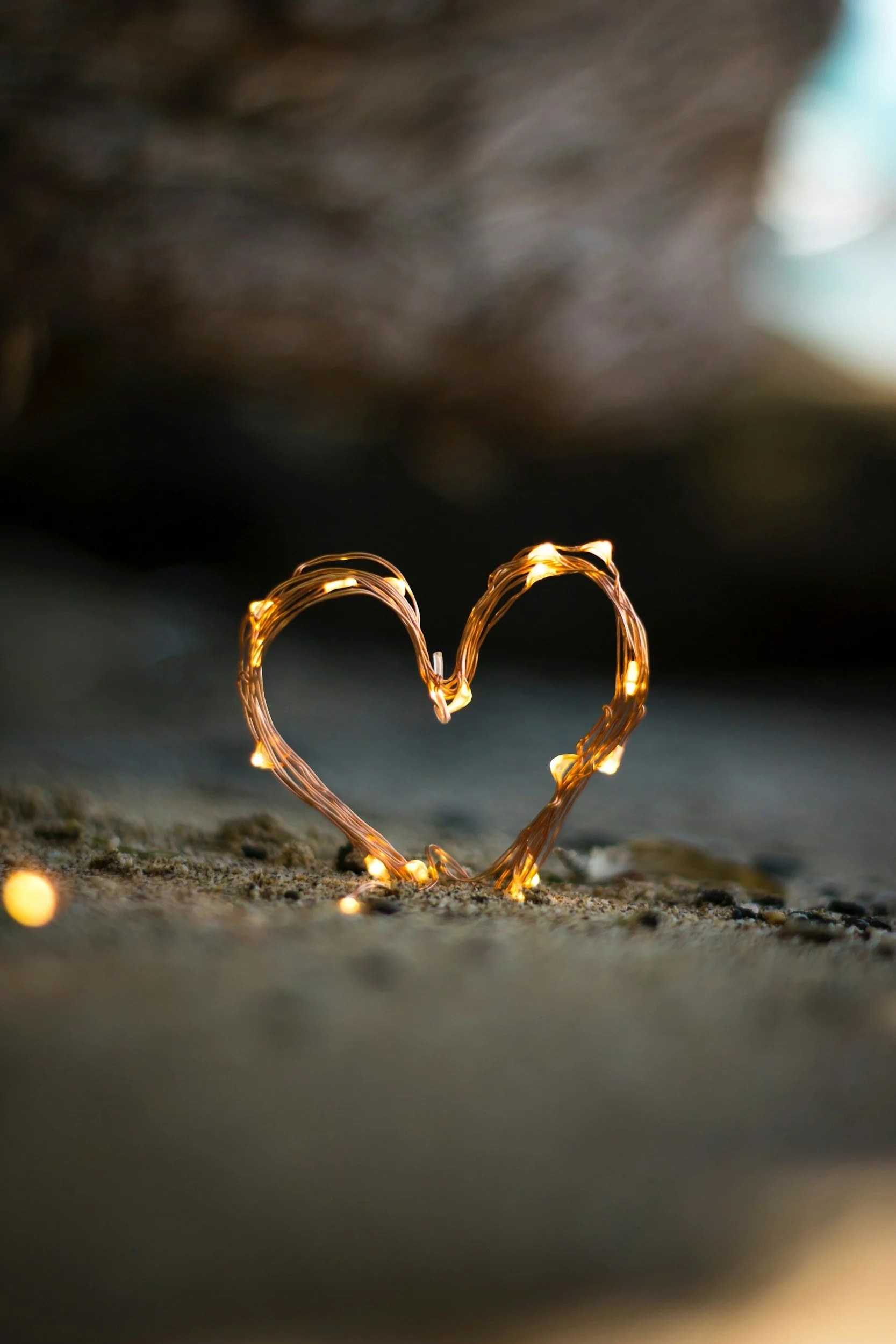 String: Heart-shaped light sculpture made of warm-colored fairy lights on a sandy surface, blurred background with cool tones.