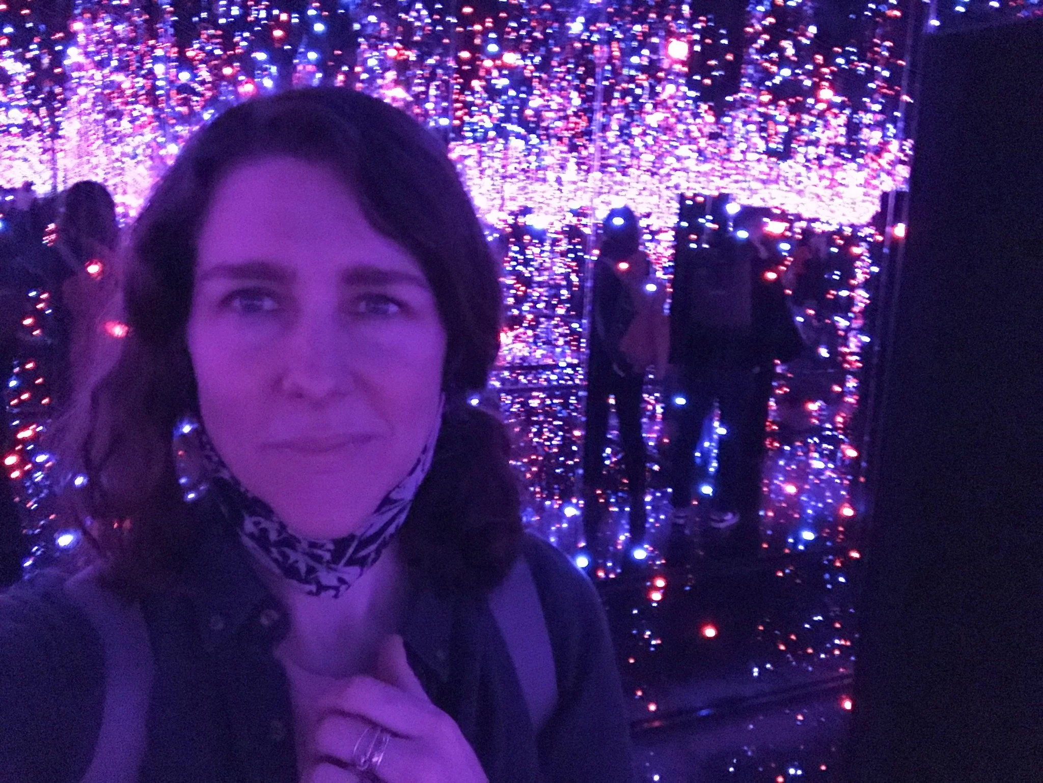 A woman taking a selfie in front of an illuminated mirror with colorful LED lights inside an exhibit or art installation.