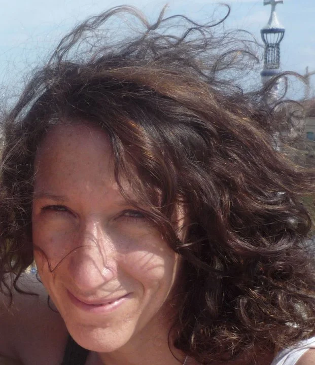 Close-up of a smiling woman with curly, wind-blown brown hair, outdoors on a sunny day, with a structure resembling a lighthouse in the background.