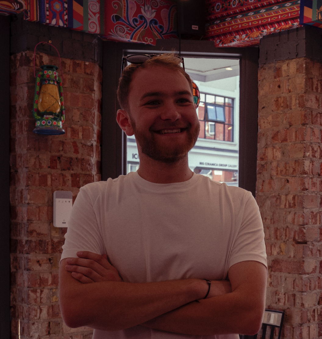 A smiling young man with crossed arms inside a restaurant with exposed brick walls, colorful Mexican-themed decorations, and a window showing buildings outside.