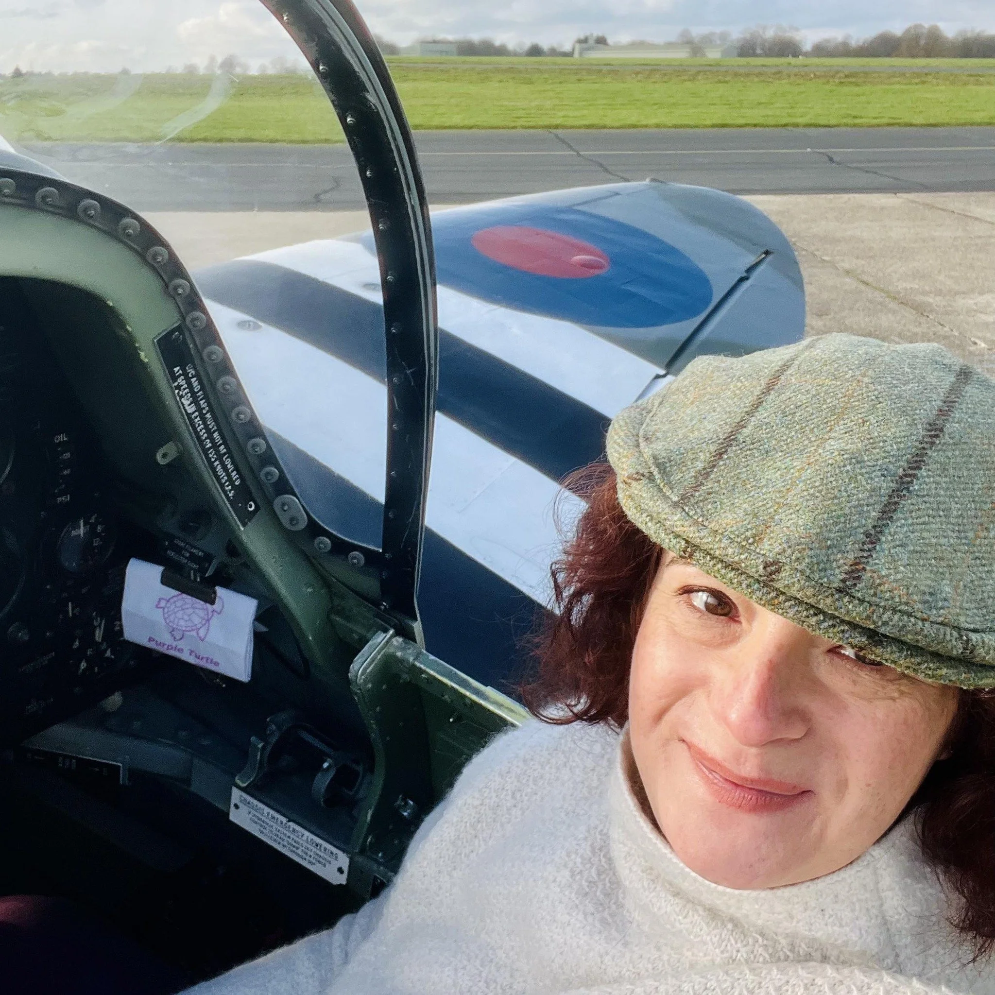 A woman in a pilot's seat taking a selfie, wearing a plaid hat, with an airplane cockpit visible behind her and a grassy field in the background.
