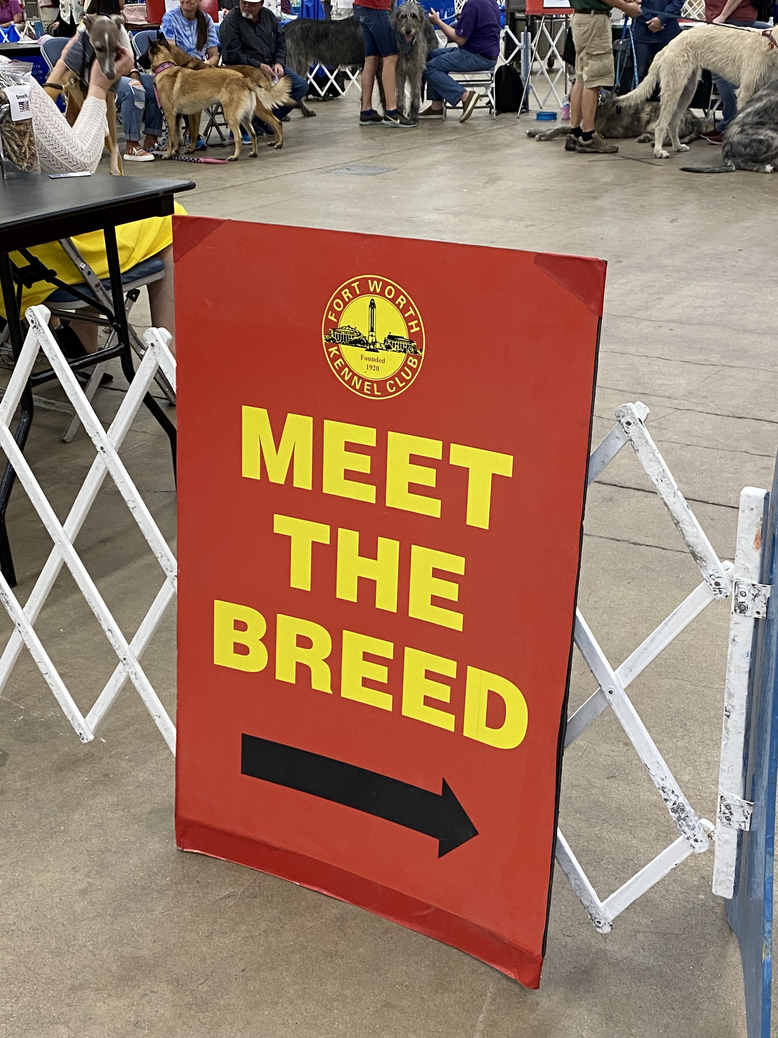 Sign for the Fort Worth Kennel Club saying 'Meet the Breed' with an arrow pointing to the right, at an indoor dog show event with people and dogs in the background.