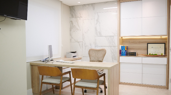 Consultório moderno e com mesa, duas cadeiras de madeira, uma cadeira de escritório ergonômica, armário branco com iluminação embutida e prateleira com livros e uma moldura da clínica DermaUro.