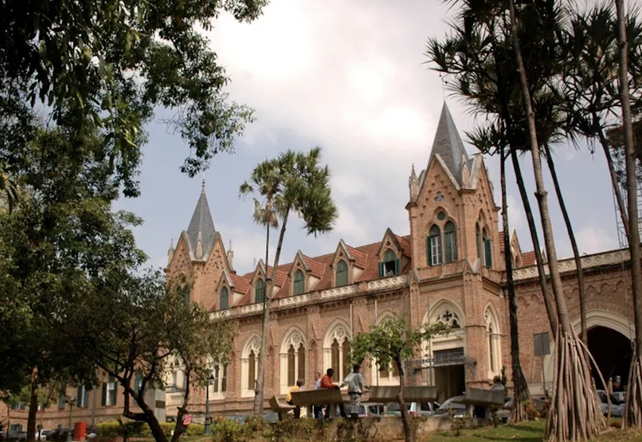 Prédio com arquitetura estilo gótico, feito de pedra vermelha, com torres pontiagudas e janelas arqueadas, rodeado de árvores e um parque com pessoas caminhando.