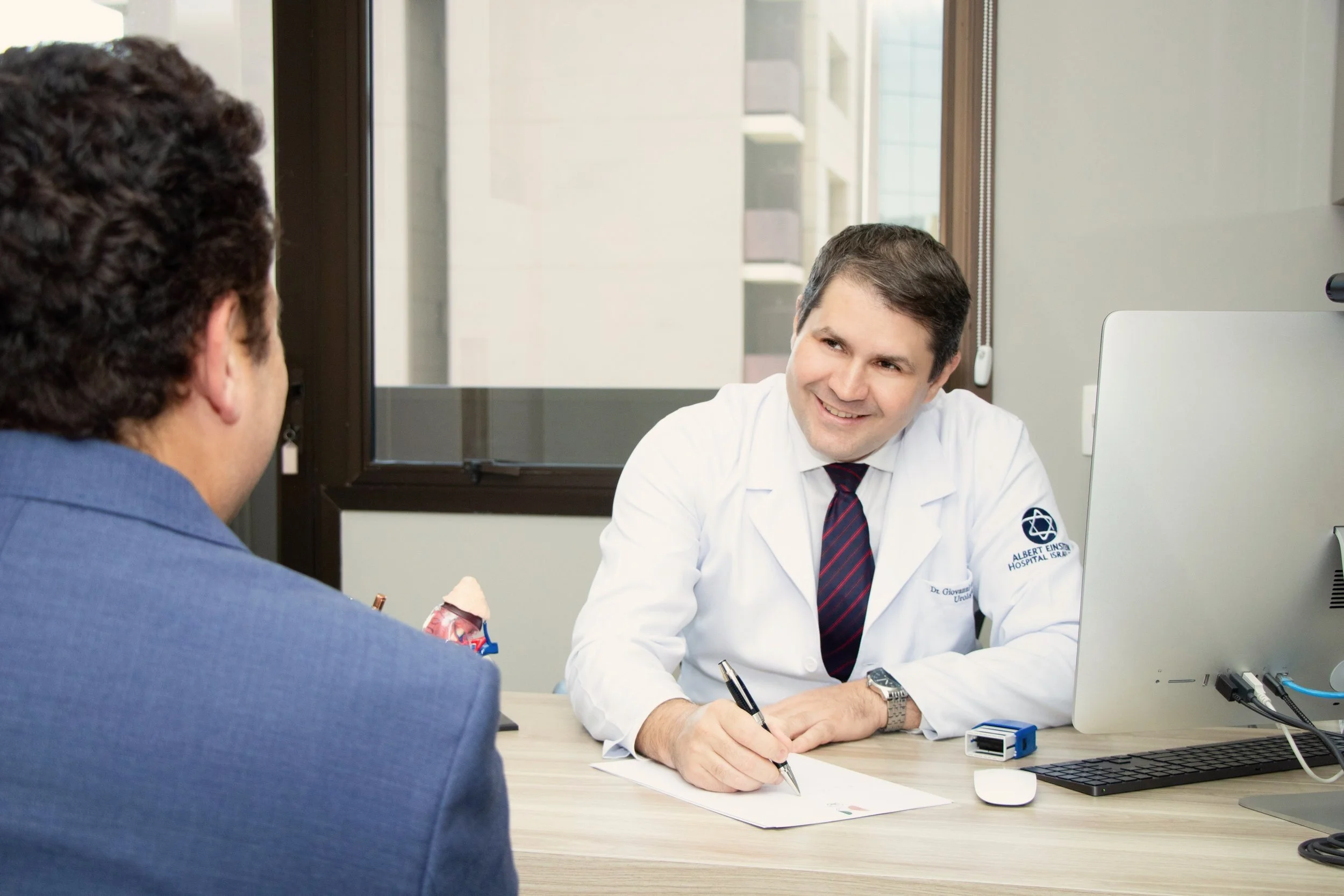 Dr. Giovanni Demartino, médico urologista, sentado na mesa conversando com um paciente, ambos olhando um para o outro. O médico está sorrindo e segurando uma caneta enquanto escreve em um papel. Há um computador e outros itens de escritório na mesa.