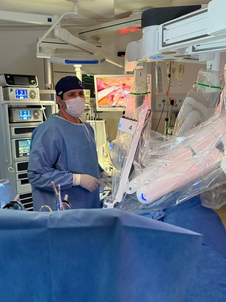 Cirurgião em sala de cirurgia usando máscara, touca e máscara, operando com equipamentos médicos e um robô cirúrgico assistente