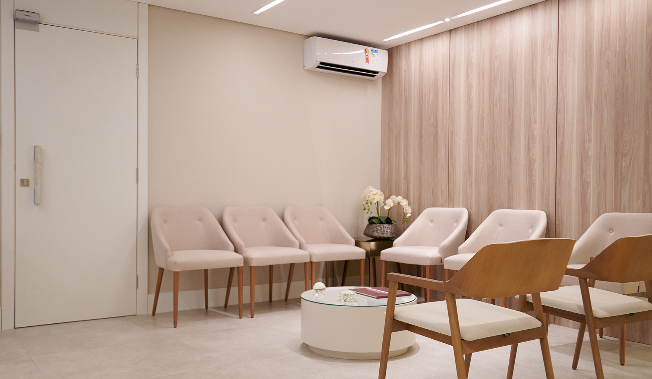 Sala de espera com cadeiras de tecido claro e madeira, mesa de centro com revistas, floricultura na parede de madeira da clínica DermaUro.