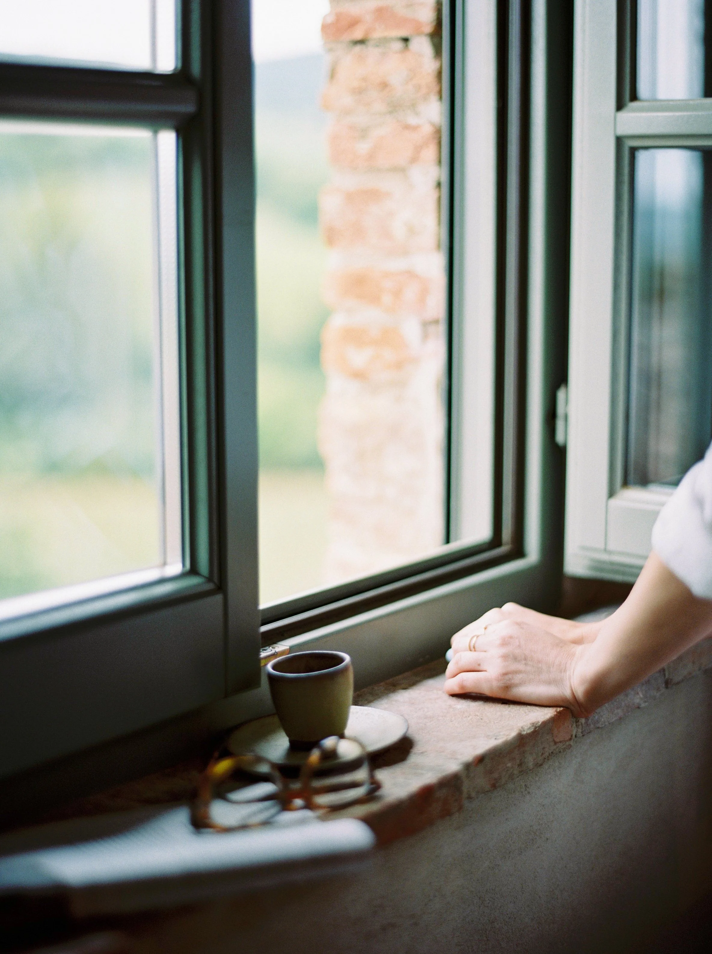 Kvinde står ved et åbent vindue med en kop kaffe og ser ud i landskabet – refleksion over relationer og værdier.