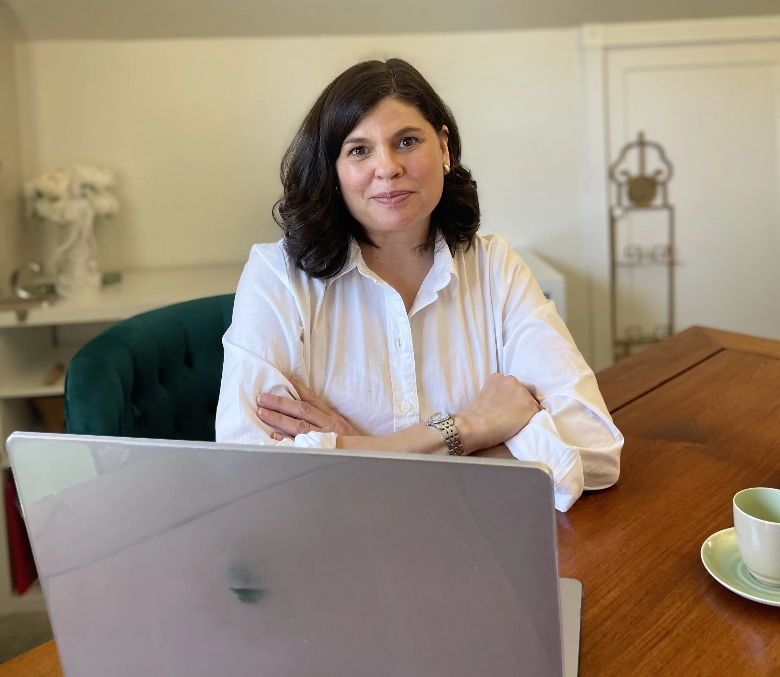 A woman with dark hair and a white shirt sitting at a wooden table with crossed arms, in front of a laptop, in a room with bookshelf and decorative items.
