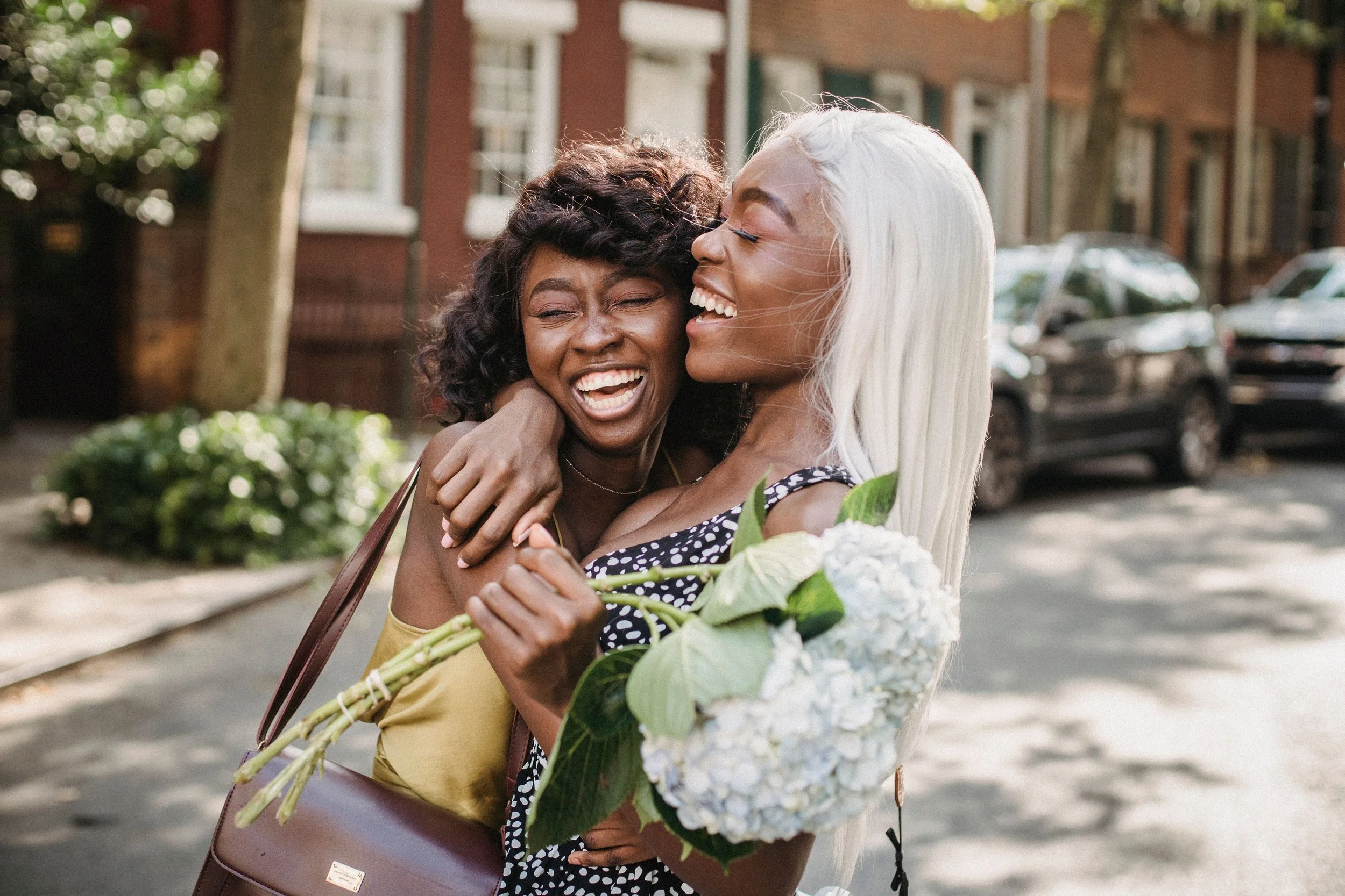 Two friends hugging and laughing outdoors while one holds a bouquet of flowers, illustrating celebration and gratitude after submitting an MBA application.