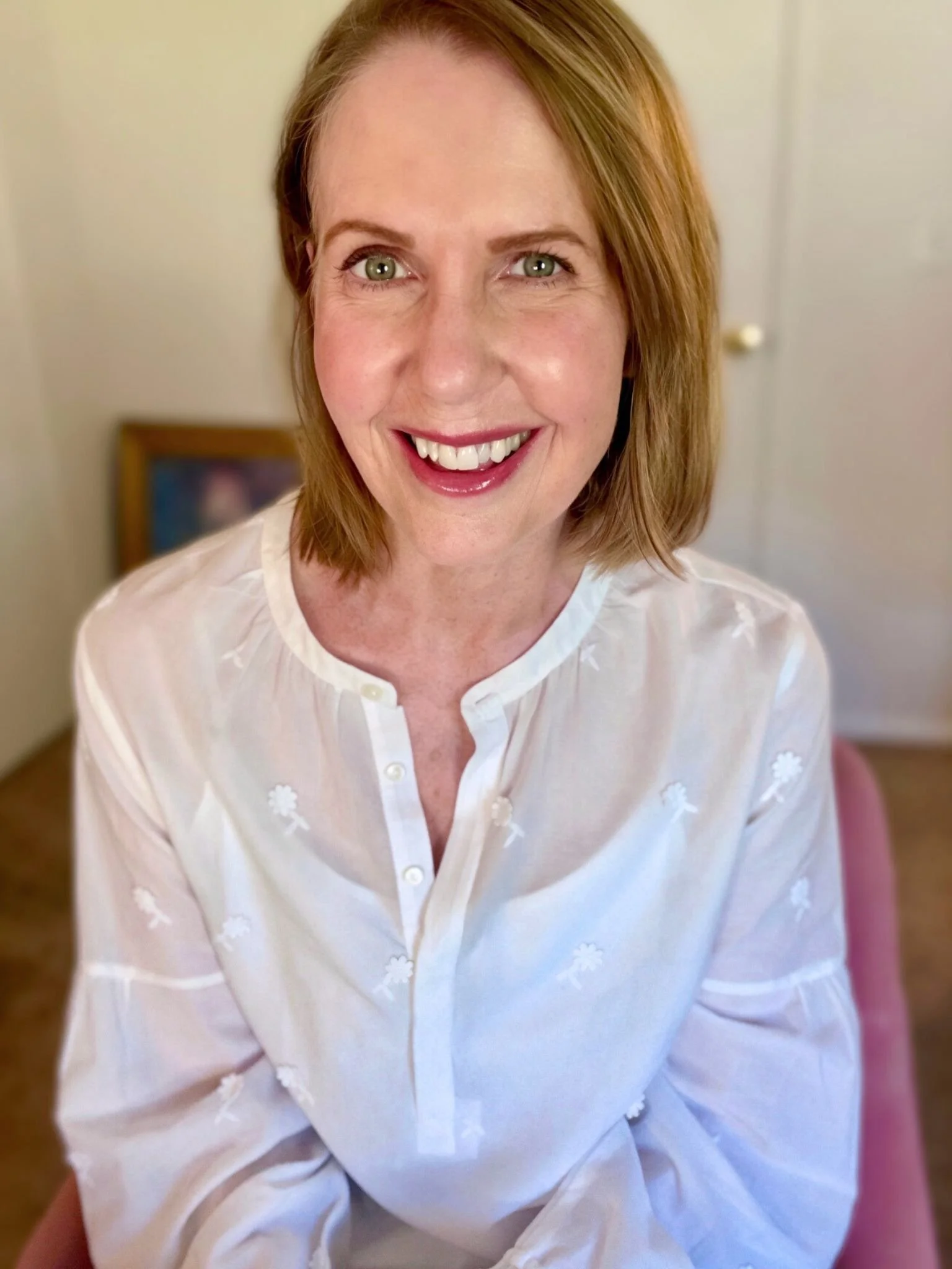 Lori Massicot sitting in a pink chair looking at the camera smiling. She is wearing a white blouse with white flowers on it.