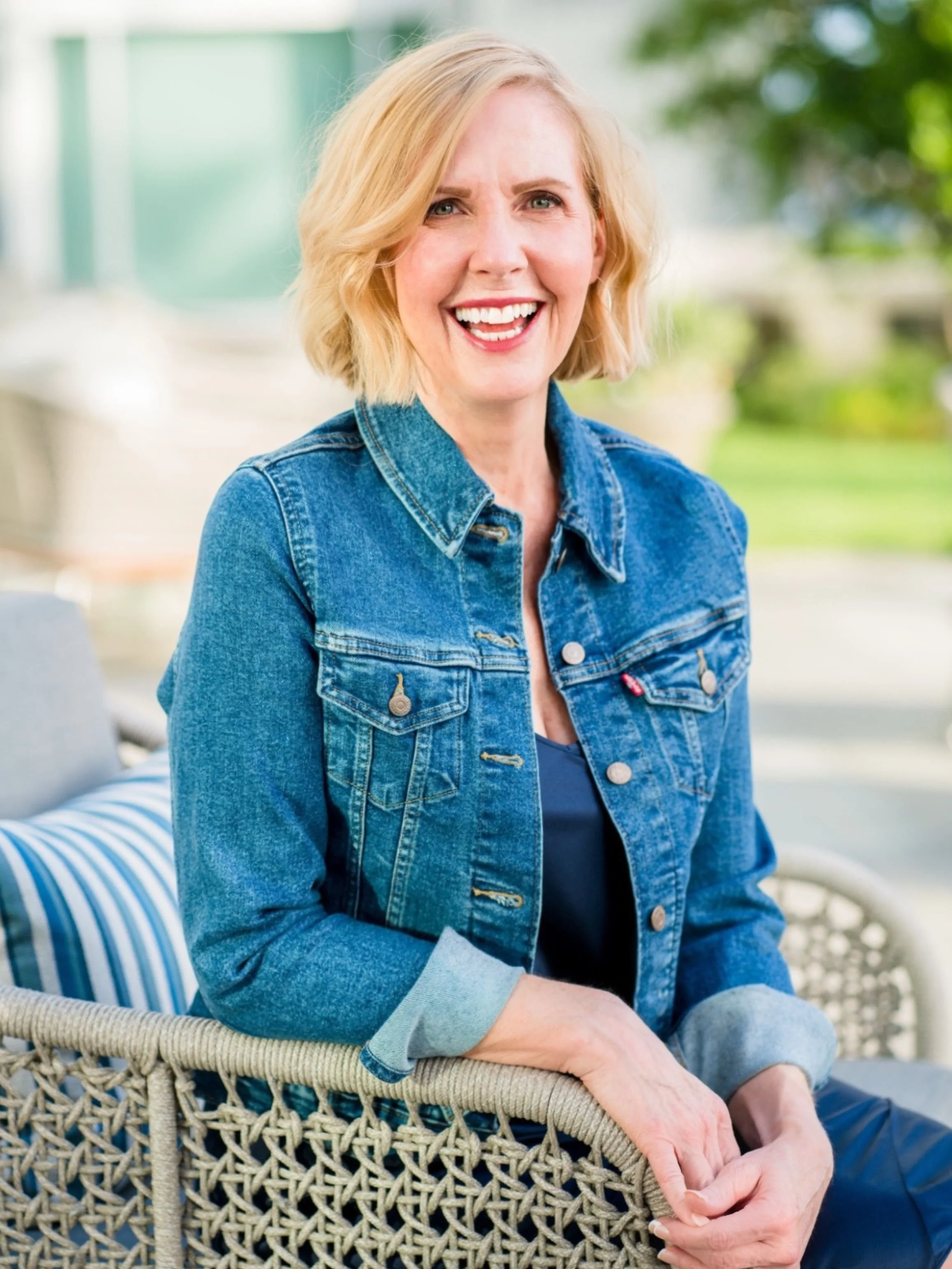 Lori Massicot smiling outdoors, wearing a denim jacket and seated on a patio chair, in a relaxed, welcoming pose.