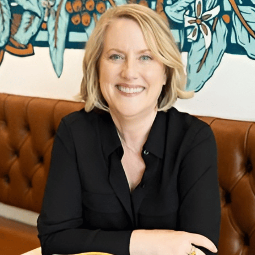 Smiling midlife woman with blonde hair wearing a black blouse, seated on a brown leather bench in front of a colorful floral mural, photographed in a relaxed, professional setting.
