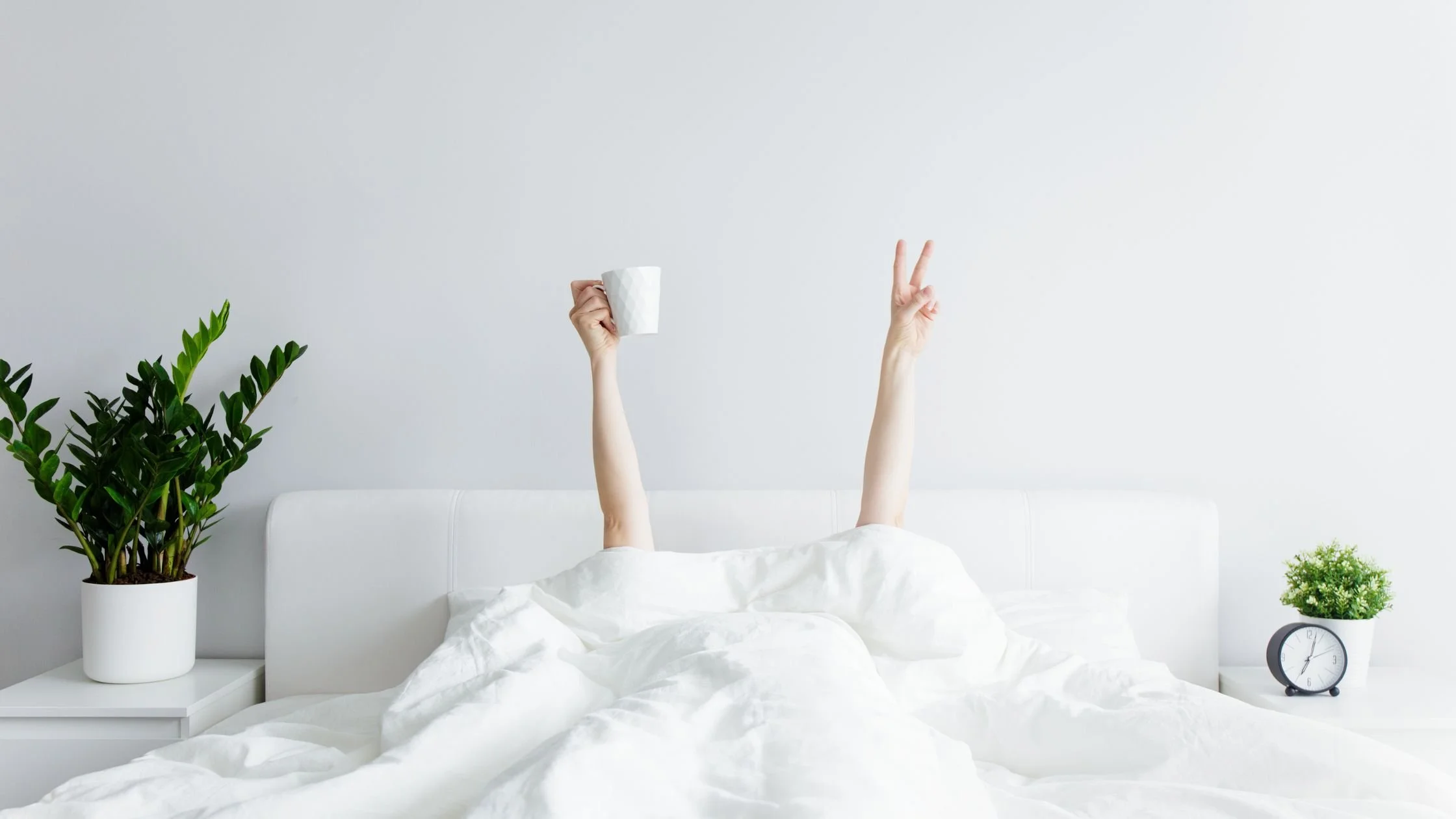 A person lying in a white bed with arms raised, holding a coffee mug in one hand and making a peace sign with the other. A small plant and alarm clock sit on a bedside table, creating a calm morning routine scene.