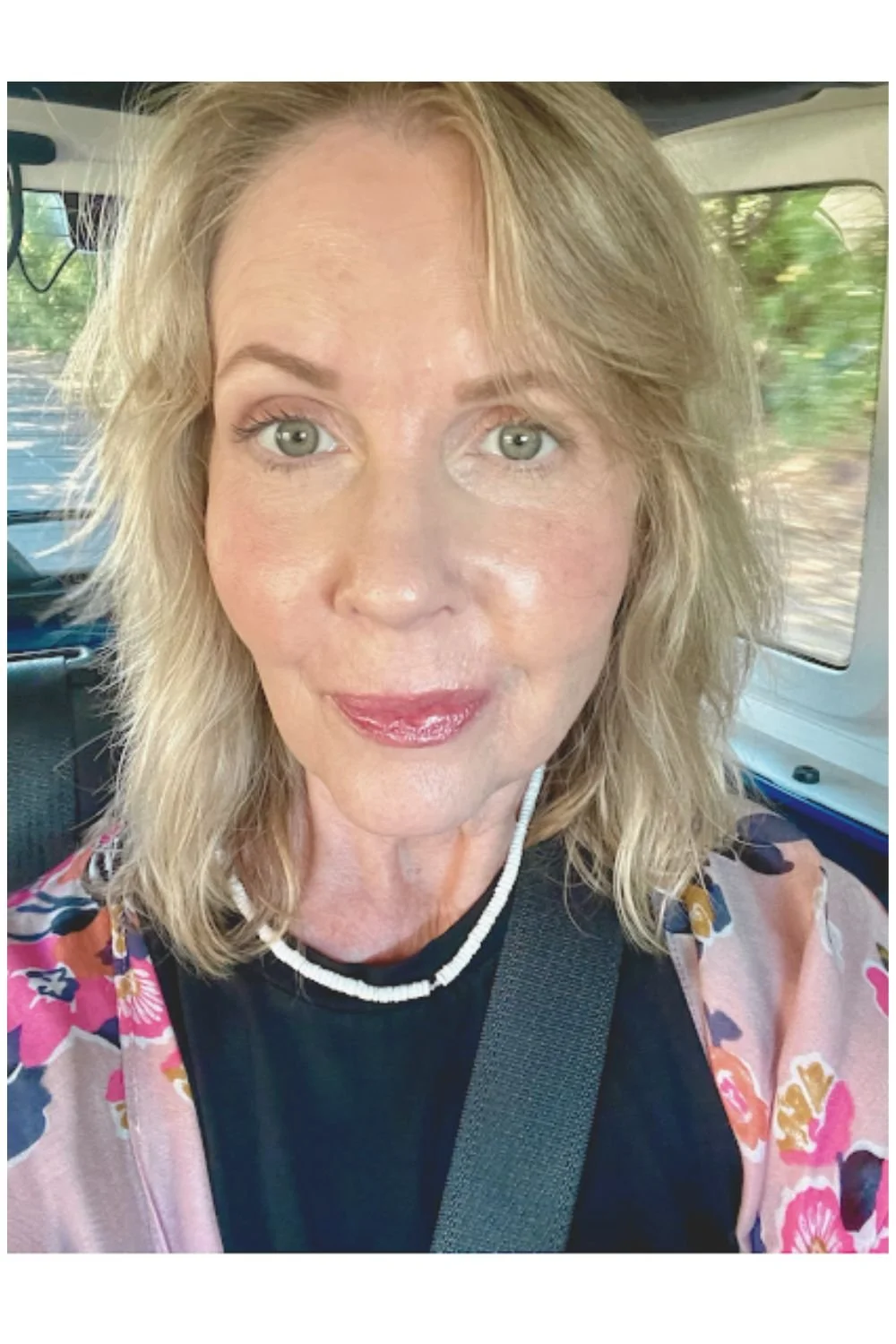 Close-up portrait of Lori Massicot sitting in a car, with shoulder-length blonde hair, green eyes, wearing a black top, a pink floral kimono-style layer, and a white beaded necklace, photographed in natural light.