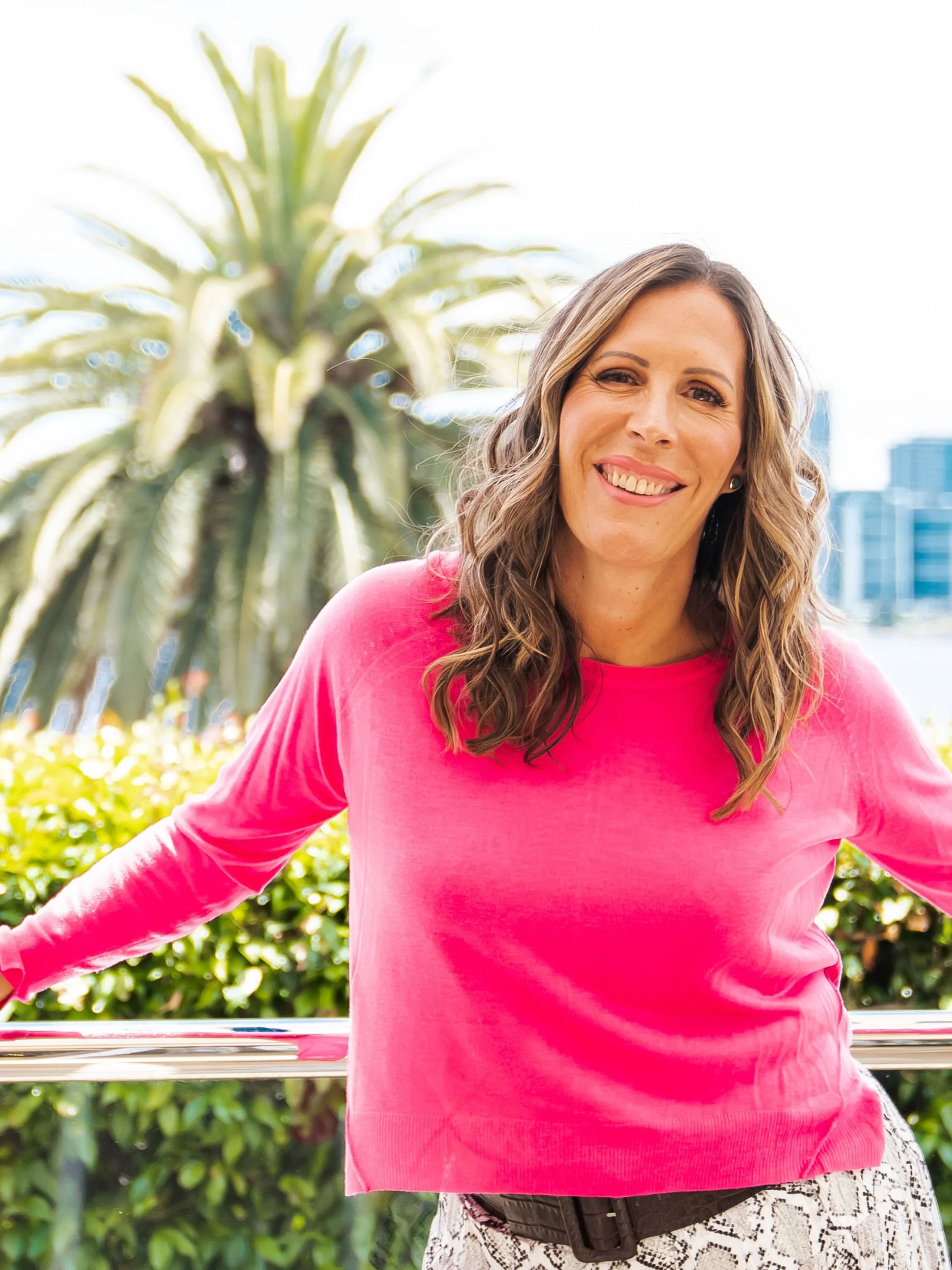 Woman in a bright pink top smiling outdoors near a palm tree with a city skyline in the background, To 50 and Beyond episode 287 cover image.