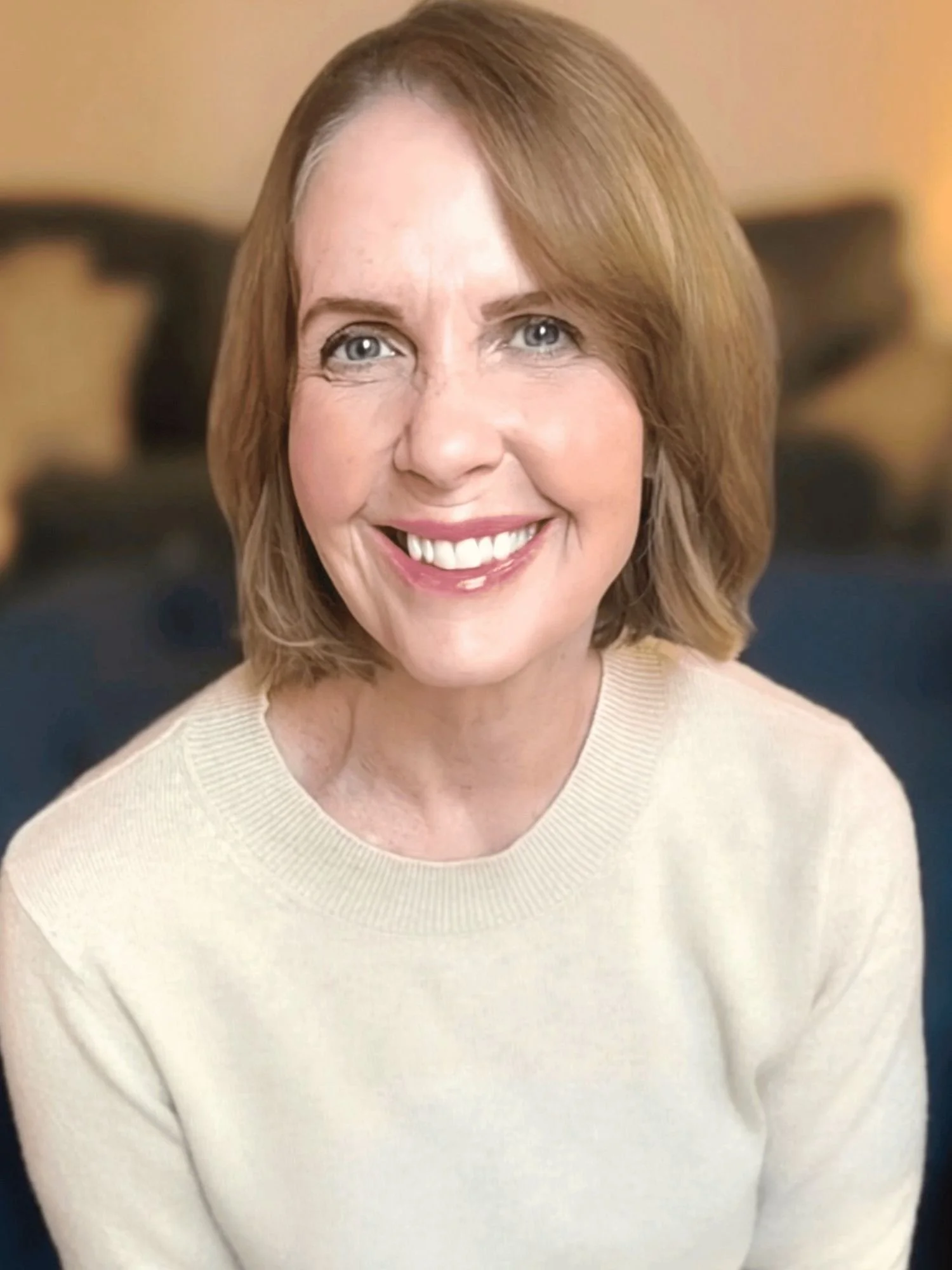 A photo of a midlife women with brownish blonde hair smiling at the camera wearing a cream colored sweater sitting in a dark blue chair.